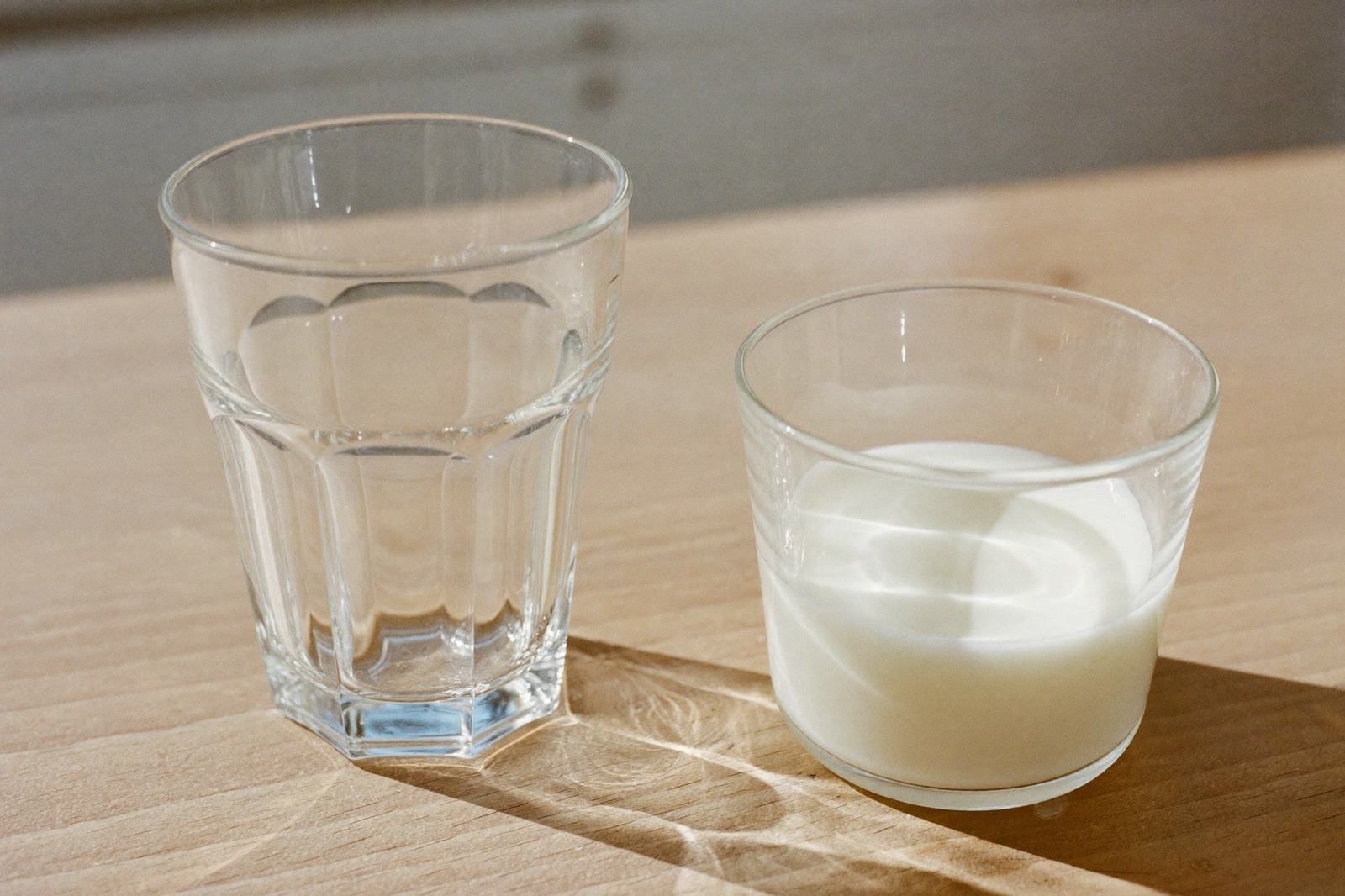 Vaso pequeño de agua y un vaso de leche sobre una mesa de madera clara con luz de mañana