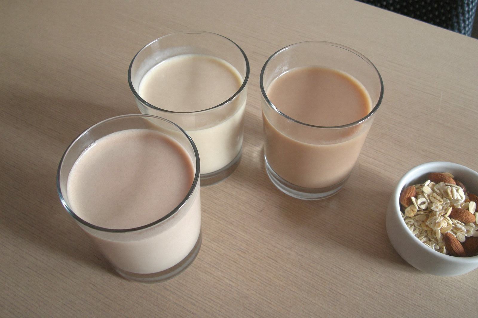 Tres vasos de vidrio con bebidas vegetales de distinto tono sobre una mesa de madera clara, con luz natural
