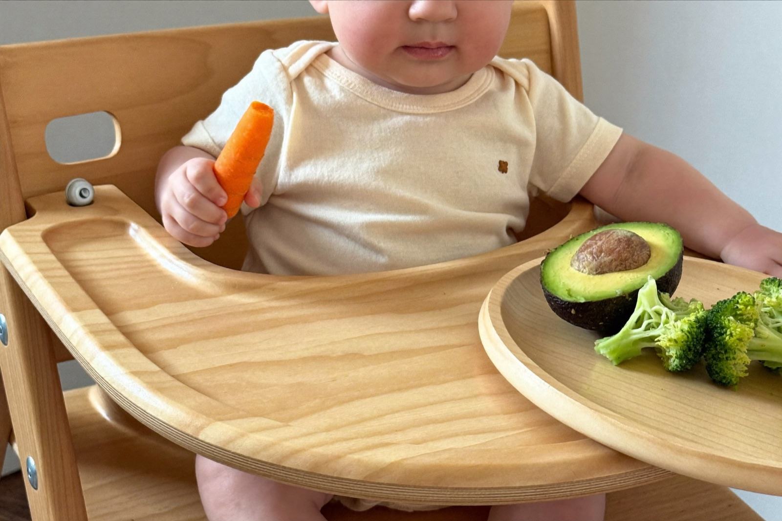 Un bebé de siete meses sentado en su periquera toma con la mano un bastón de zanahoria cocida, con aguacate y brócoli en la charola