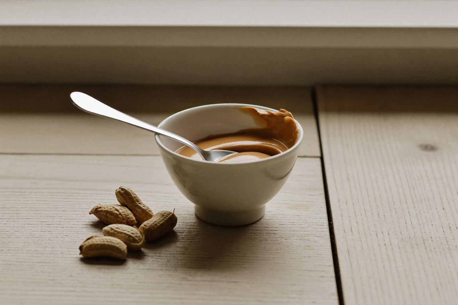 Un tazón pequeño con crema de cacahuate y una cuchara sobre una mesa de madera clara, con cacahuates enteros al lado