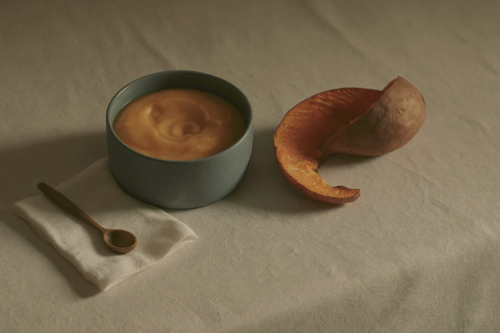 Tazón bajo de cerámica mate azul verdoso con puré de camote de superficie lisa, una cuchara pequeña de madera sobre una servilleta de lino doblada y un trozo de camote asado con la cáscara enrollada, todo sobre lino color crema con luz cálida