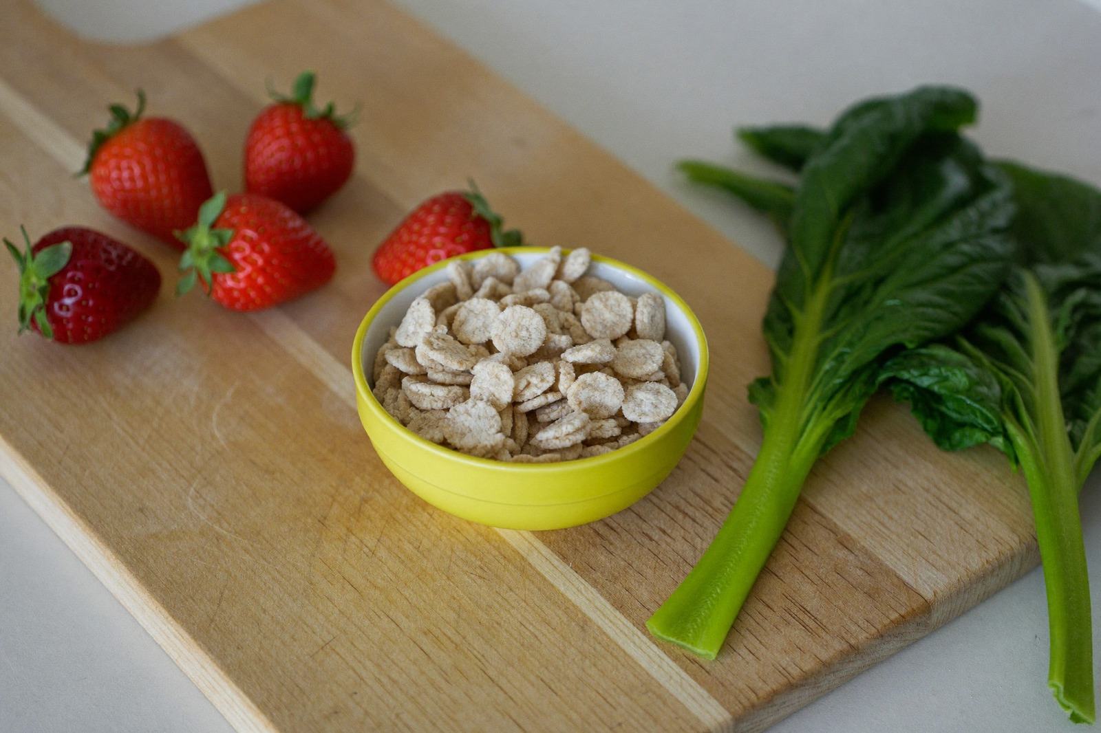 Tazón de cereal infantil junto a fresas y hojas verdes frescas sobre una tabla de madera clara