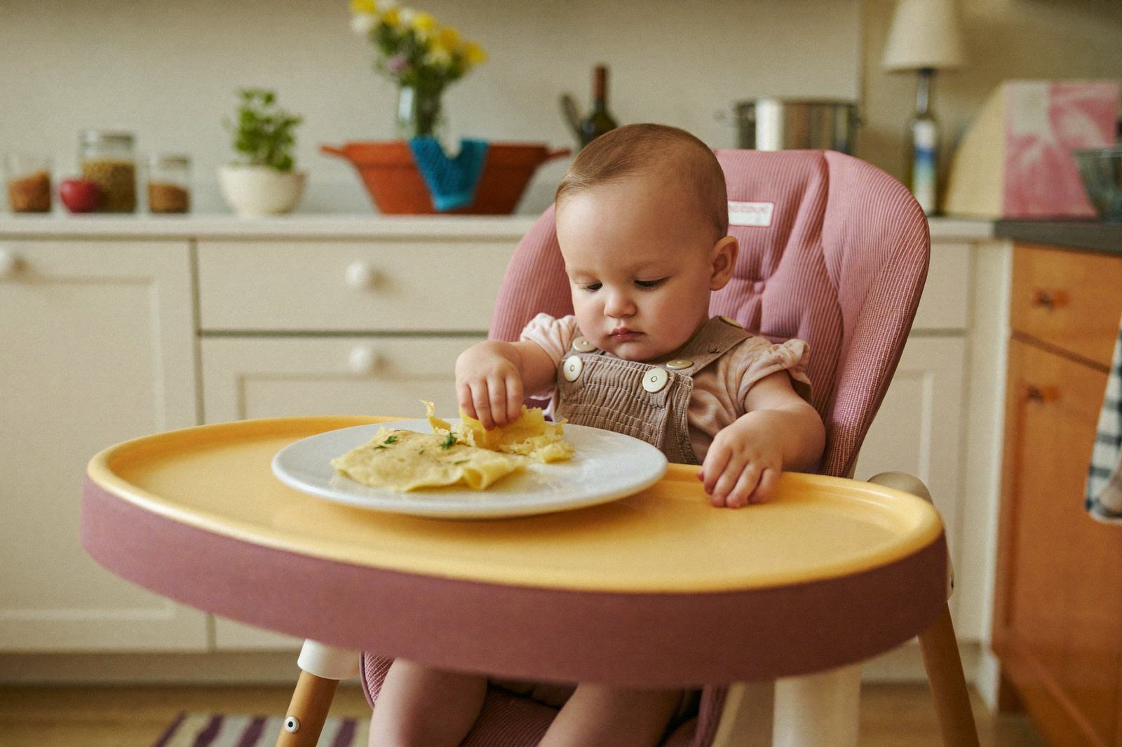 Bebé en silla alta frente a un plato con trozos de comida, tomando uno con la mano
