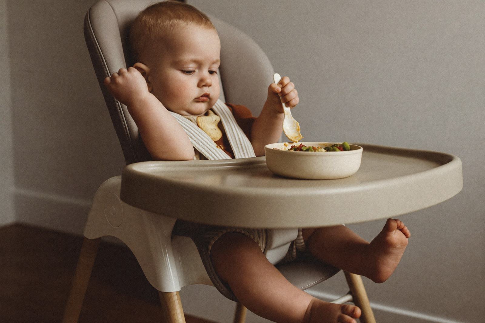 Bebé en silla alta sosteniendo una cuchara pequeña con el puño sobre una bandeja con comida