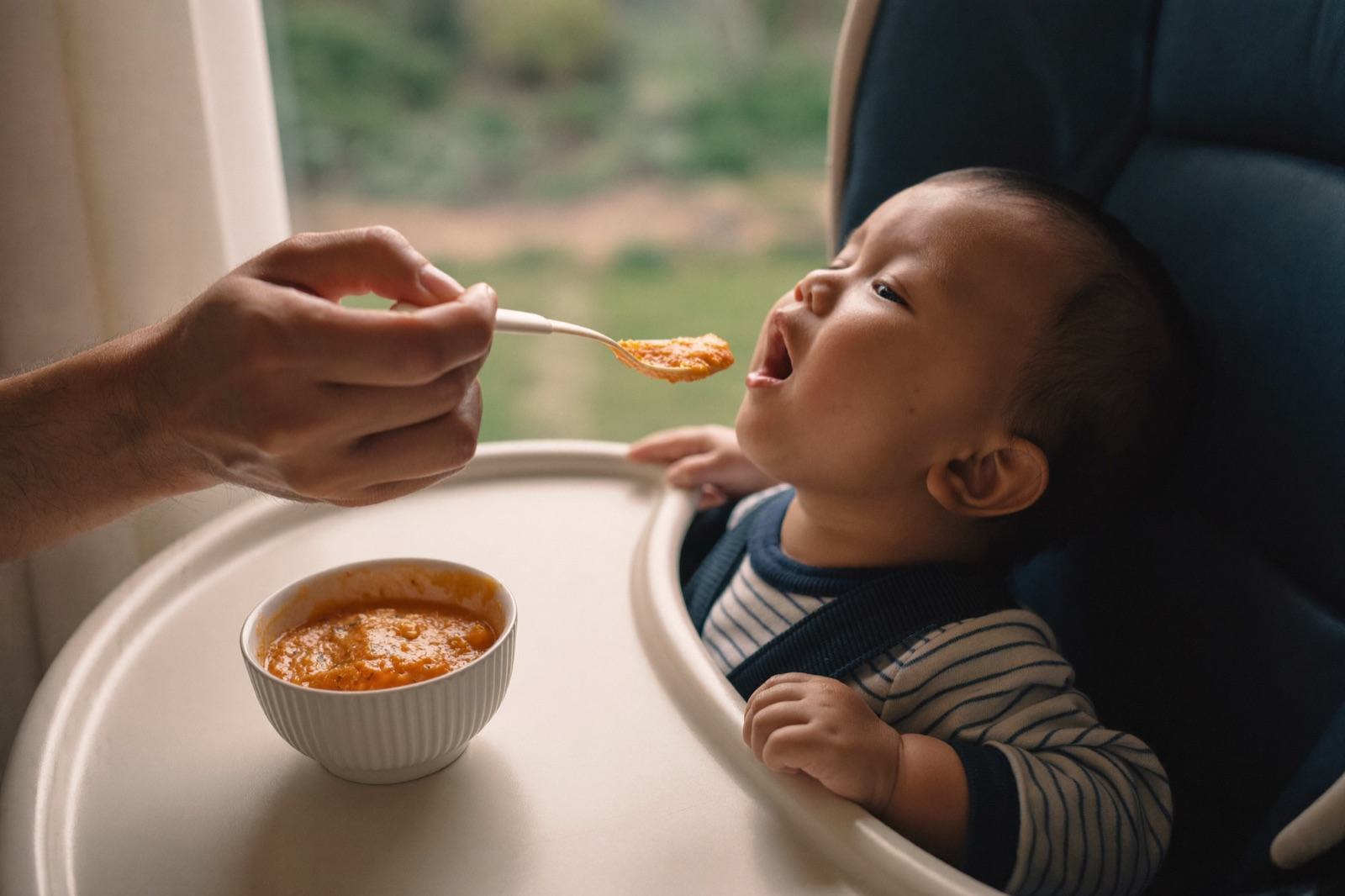 La mano de un papá acerca una cucharita con puré de verdura a un bebé sentado en la periquera, que abre la boca
