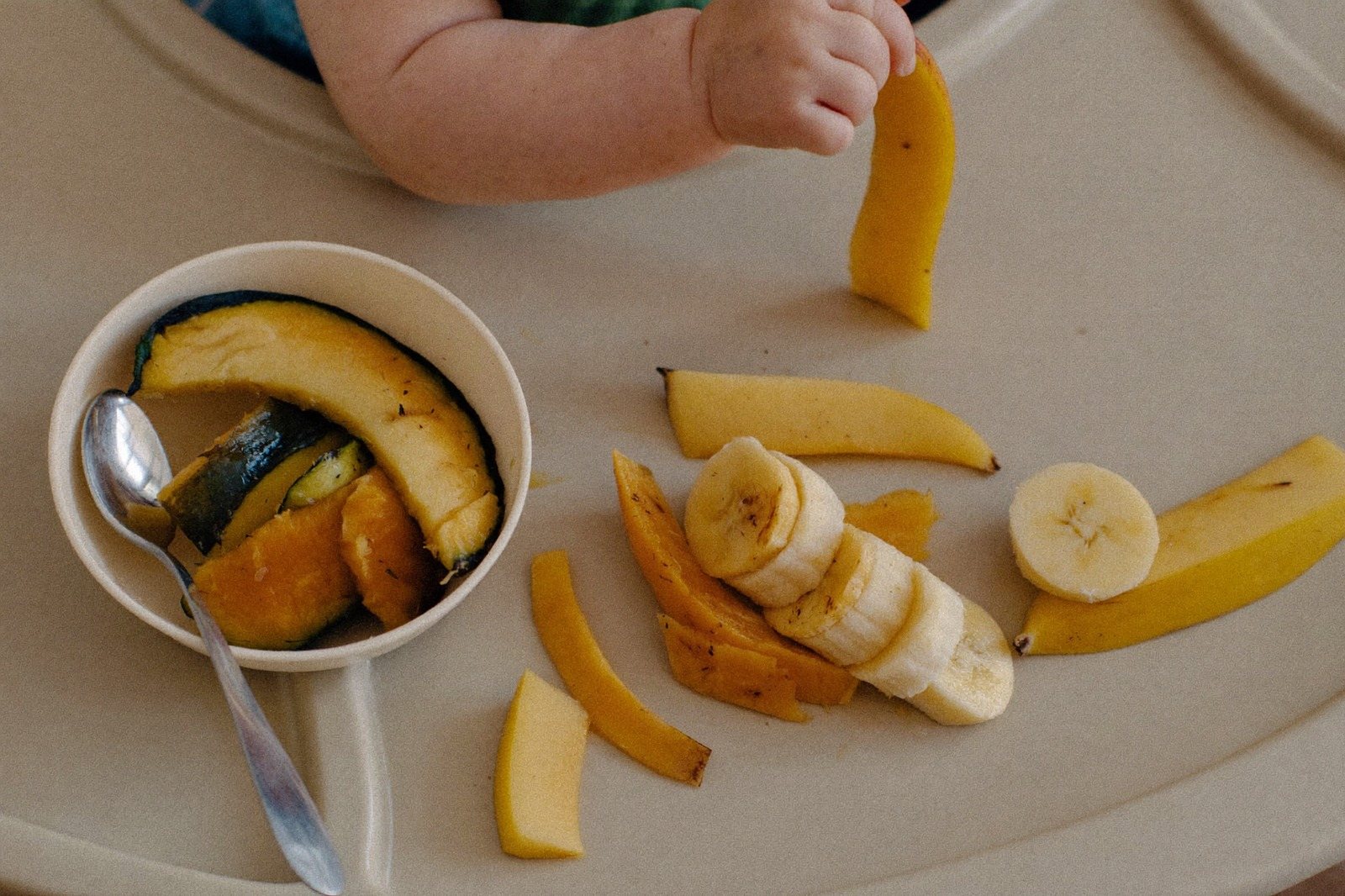 Bandeja de silla alta con bastones de calabaza y plátano cortados en tiras, y una mano de bebé tomando uno