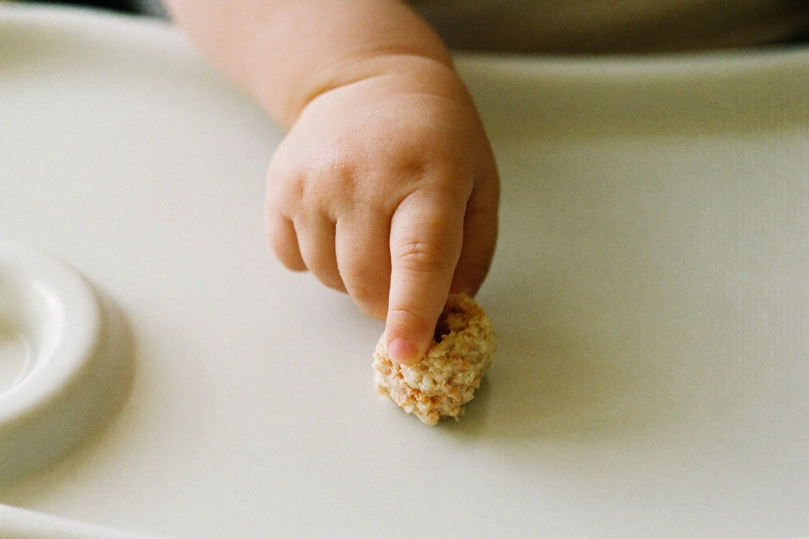 Mano de bebé tomando un trozo pequeño de comida con el pulgar y el índice sobre una bandeja clara