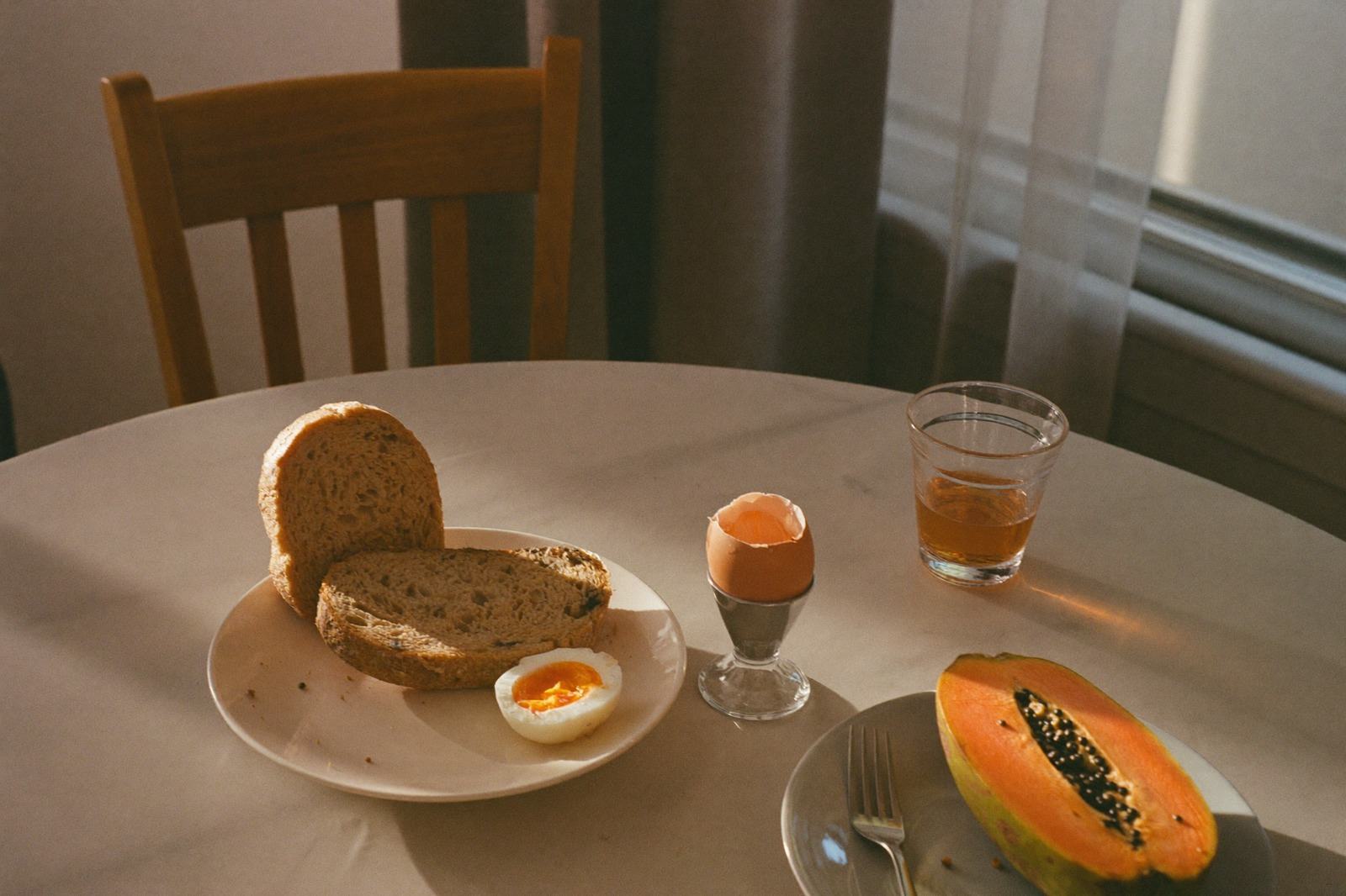 Mesa de desayuno con pan integral, huevo tibio, papaya y un vaso, junto a una silla de madera