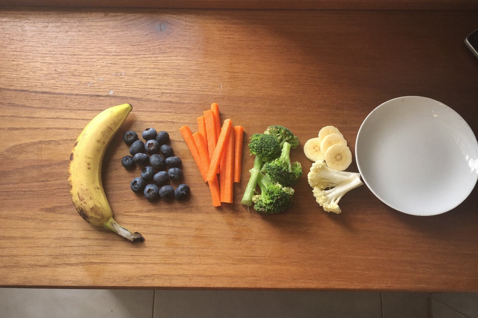 Cinco porciones de frutas y verduras acomodadas sobre una mesa de madera junto a un tazón blanco