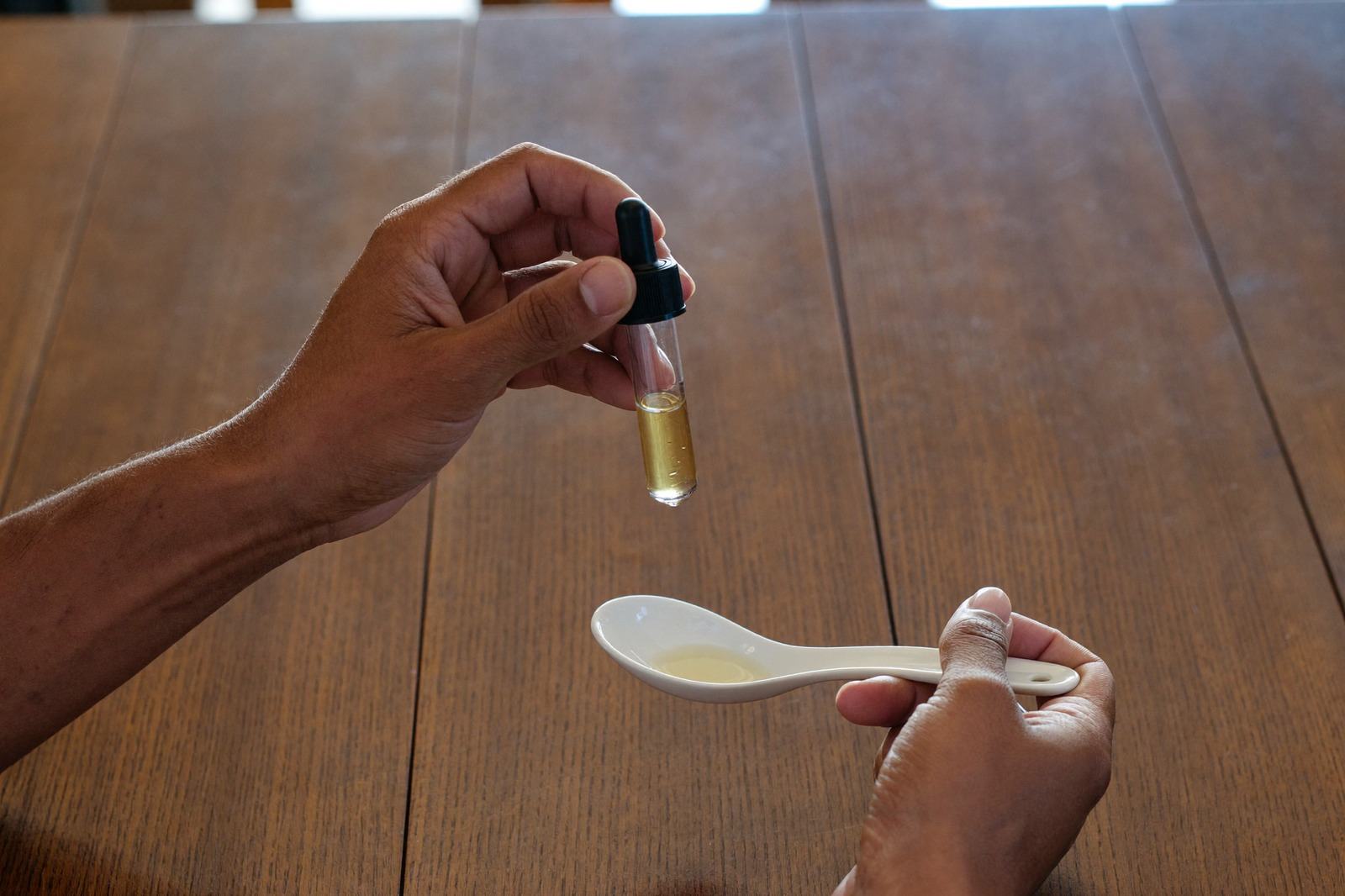 Manos sosteniendo un gotero de vidrio sobre una cuchara blanca, en una mesa de madera