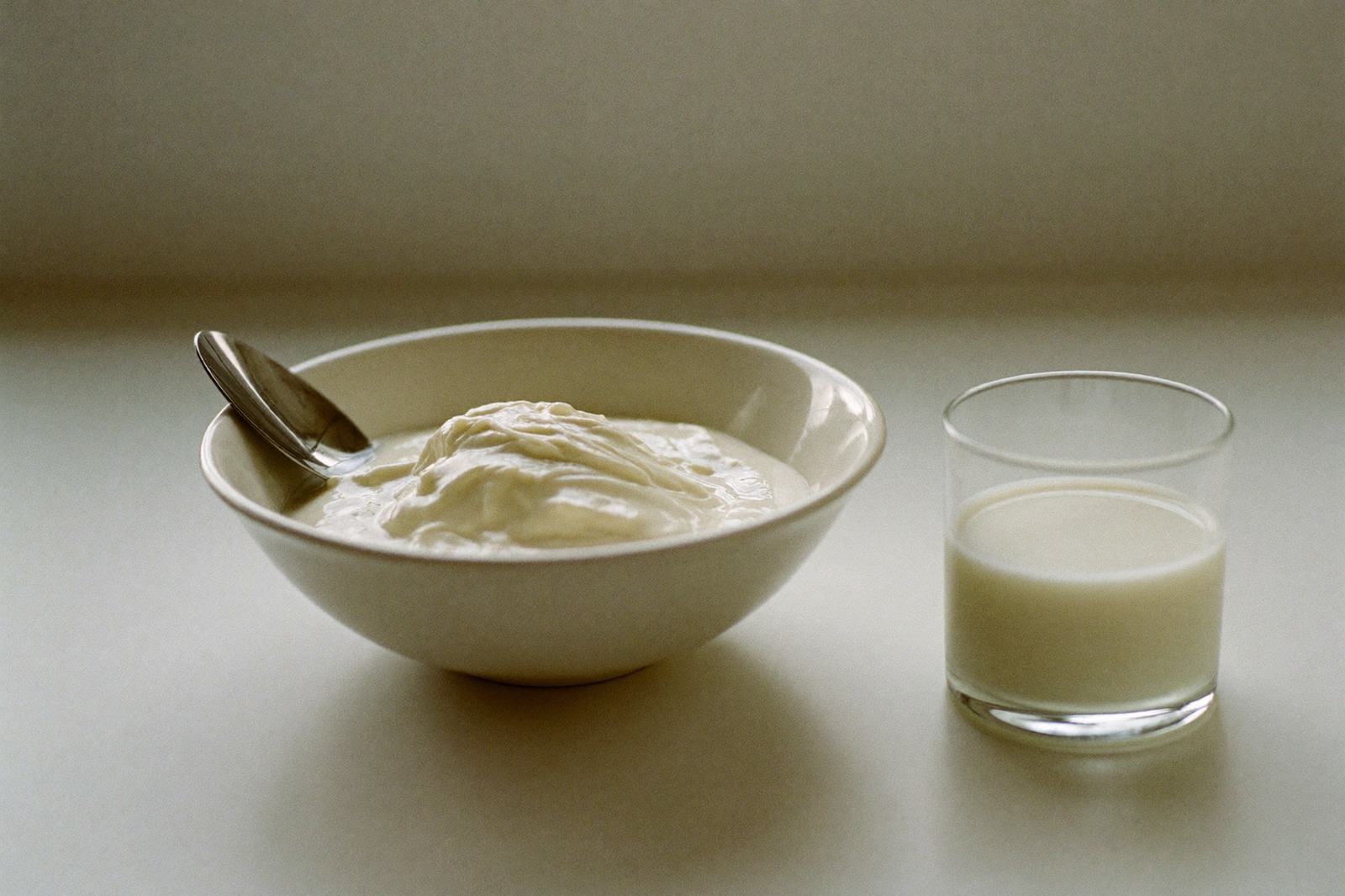 Tazón pequeño de yogur natural con una cuchara, sobre una mesa de madera clara junto a un vaso de leche