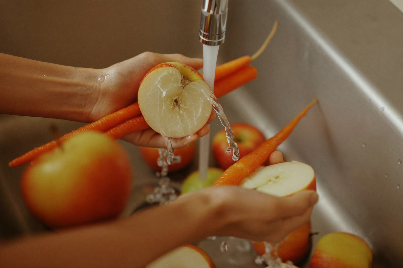 Manos lavando manzanas y zanahorias bajo el chorro del fregadero