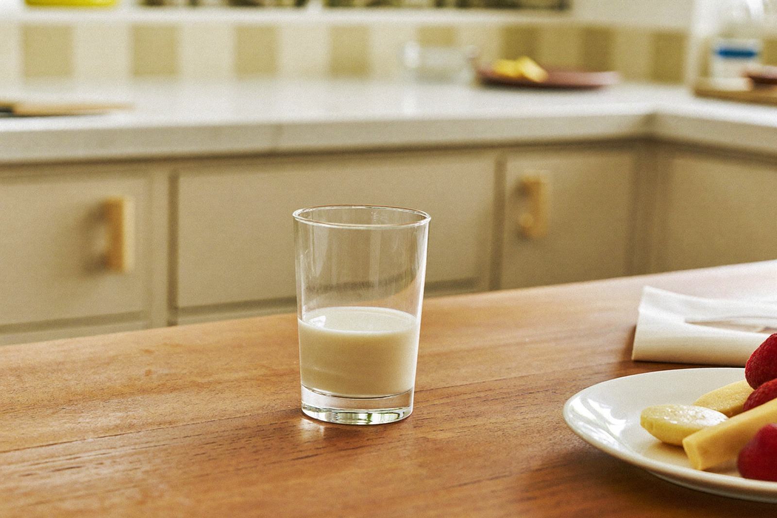 Vaso pequeño de vidrio con leche sobre una mesa de madera junto a un plato con fruta cortada