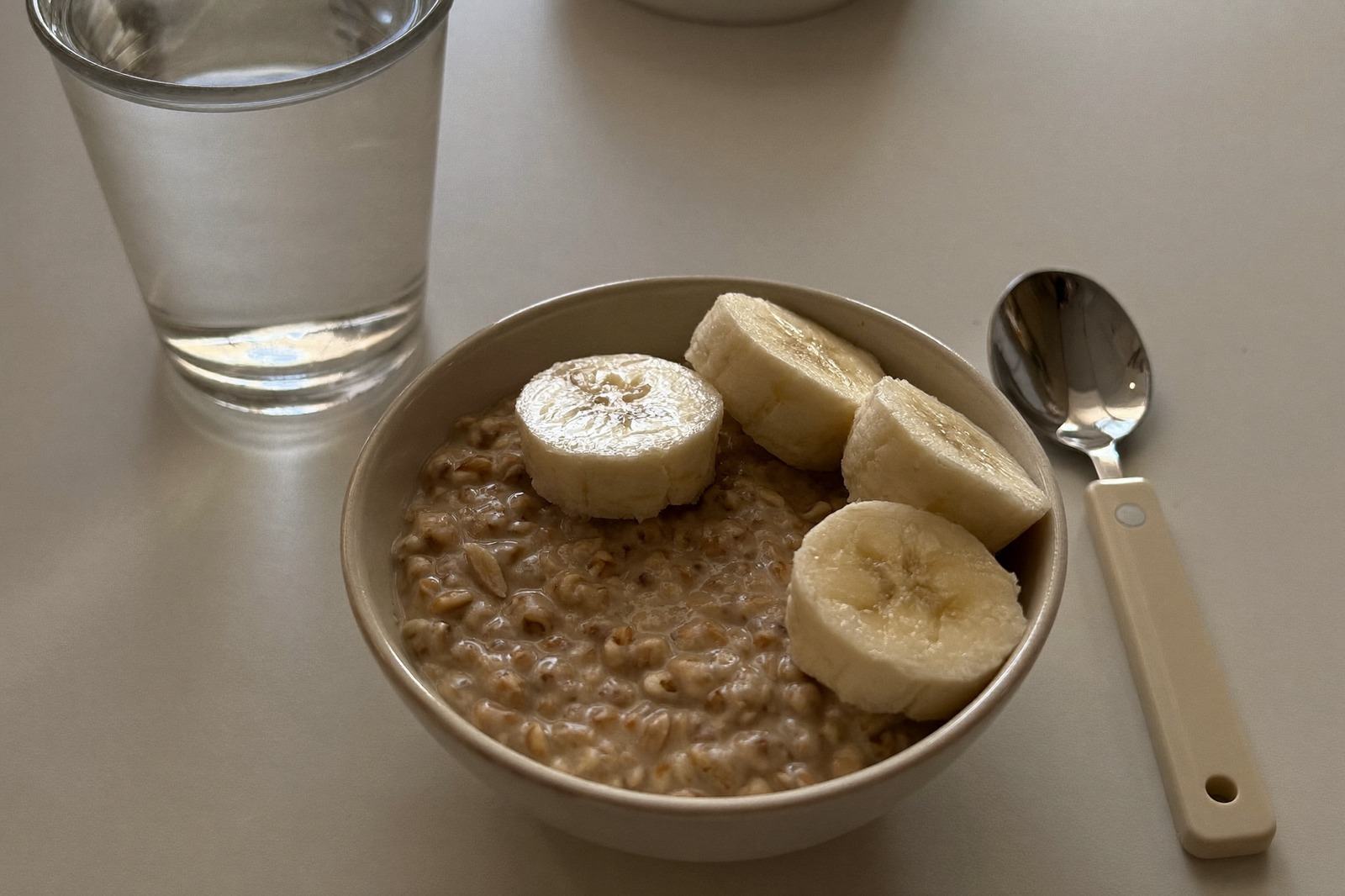 Tazón pequeño con avena, un plátano rebanado y un vaso de agua sobre una mesa clara de mañana