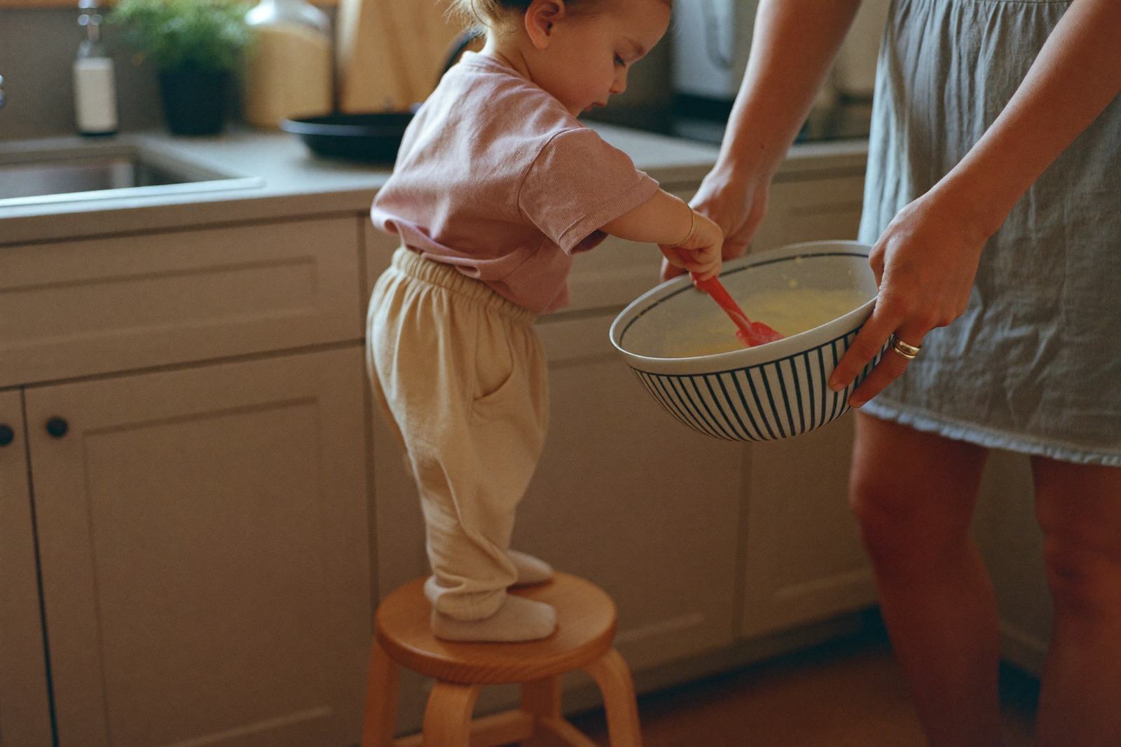 Niño pequeño de pie sobre un banquito revolviendo un tazón en la cocina junto a un adulto