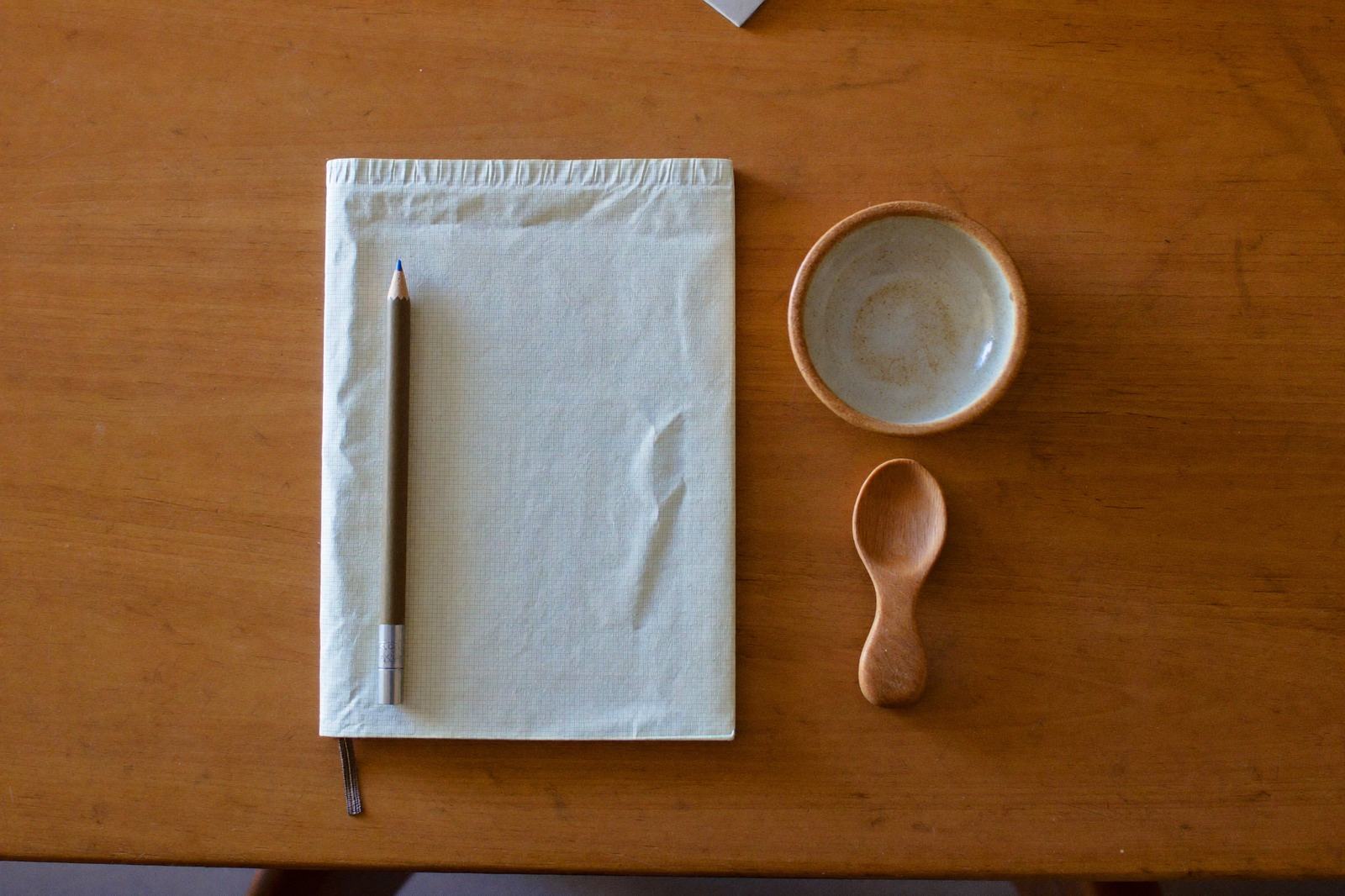Libreta cerrada, lápiz, tazón pequeño y cuchara de bebé sobre una mesa de madera clara