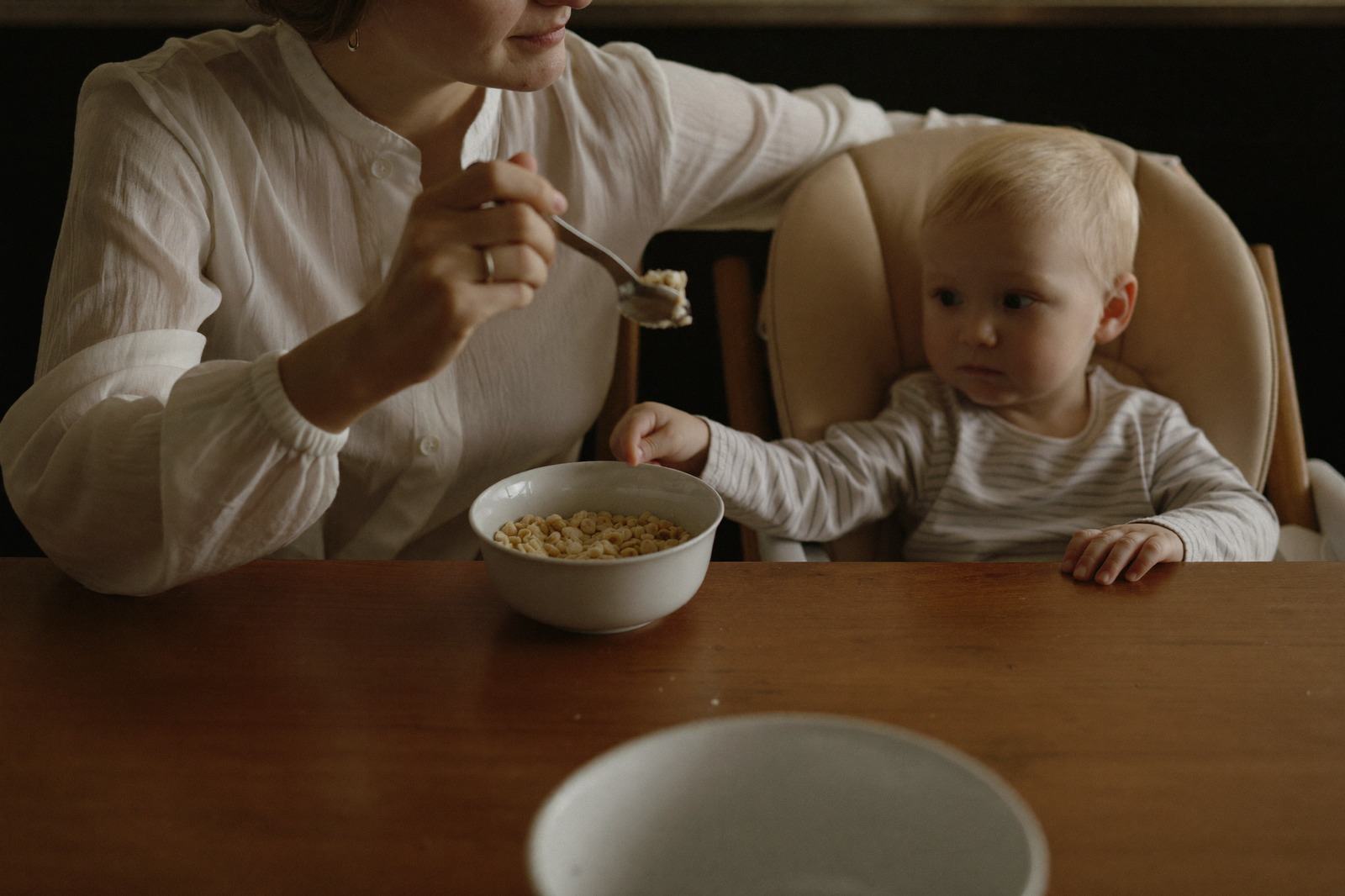 Bebé de seis meses en su silla alta frente a un tazón pequeño de cereal, con la mamá a su lado