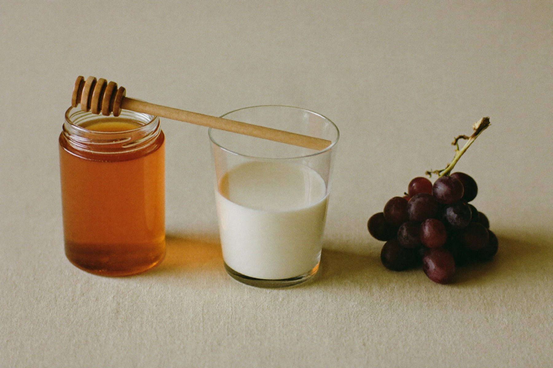 Un frasco de vidrio con miel y su cucharón, un vaso de leche entera y un racimo de uvas enteras sobre lino crudo