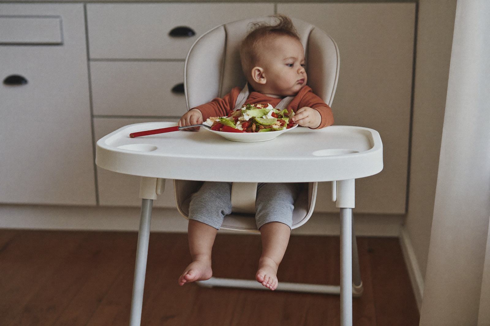 Bebé en silla alta con el plato lleno frente a él y la cara girada hacia un lado
