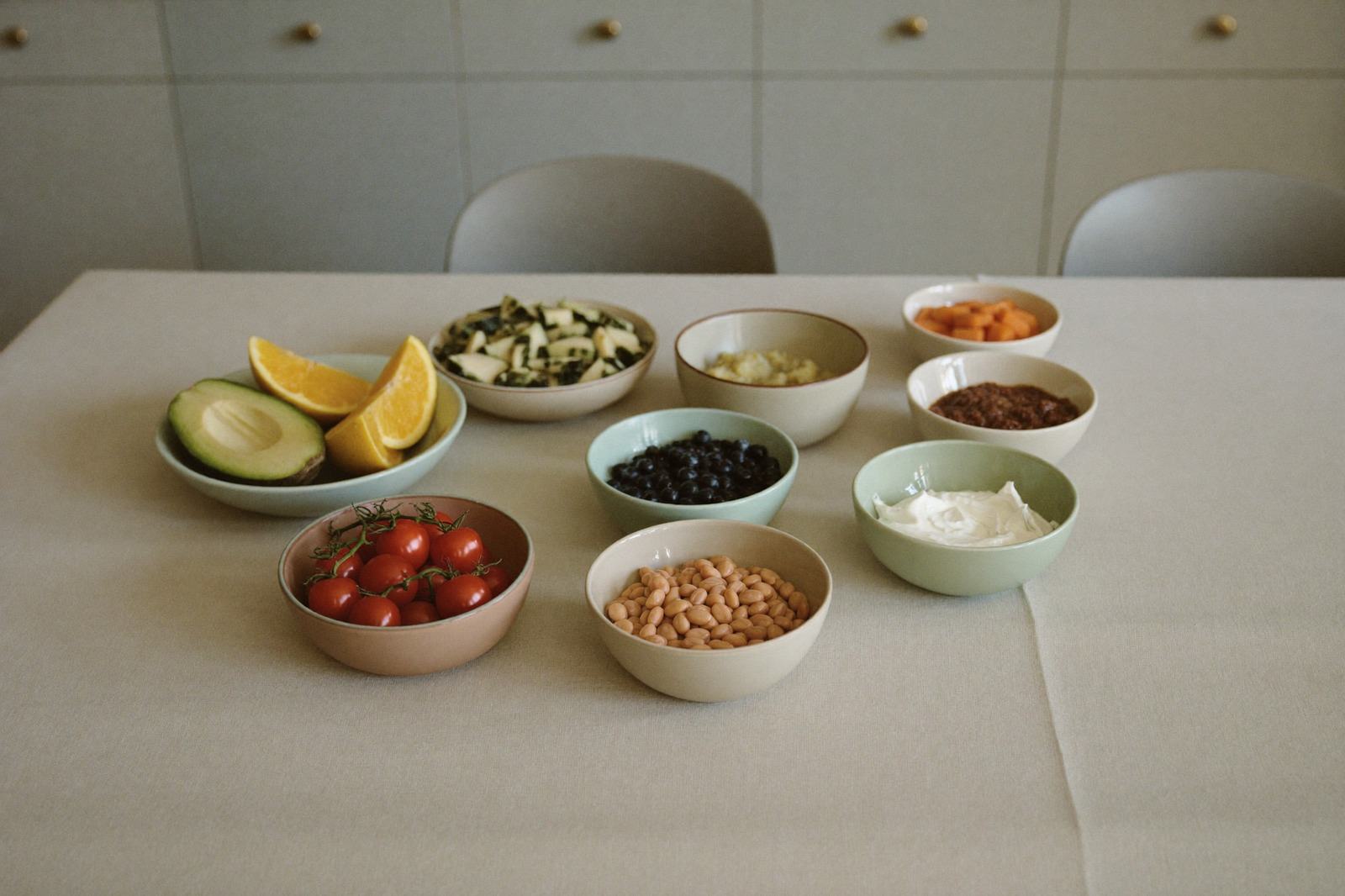 Tazones pequeños alineados sobre una mesa clara con verdura, fruta, frijol cocido y yogur natural