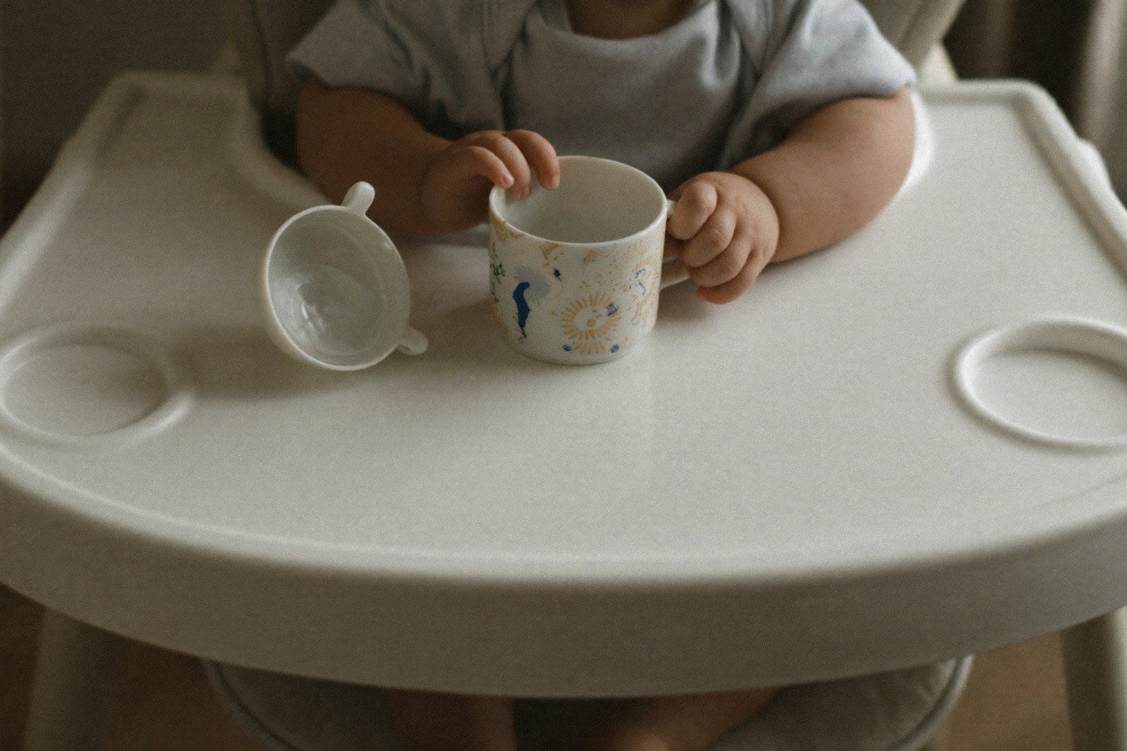 Manos de bebé sosteniendo un vaso pequeño sin tapa sobre la bandeja de una silla alta