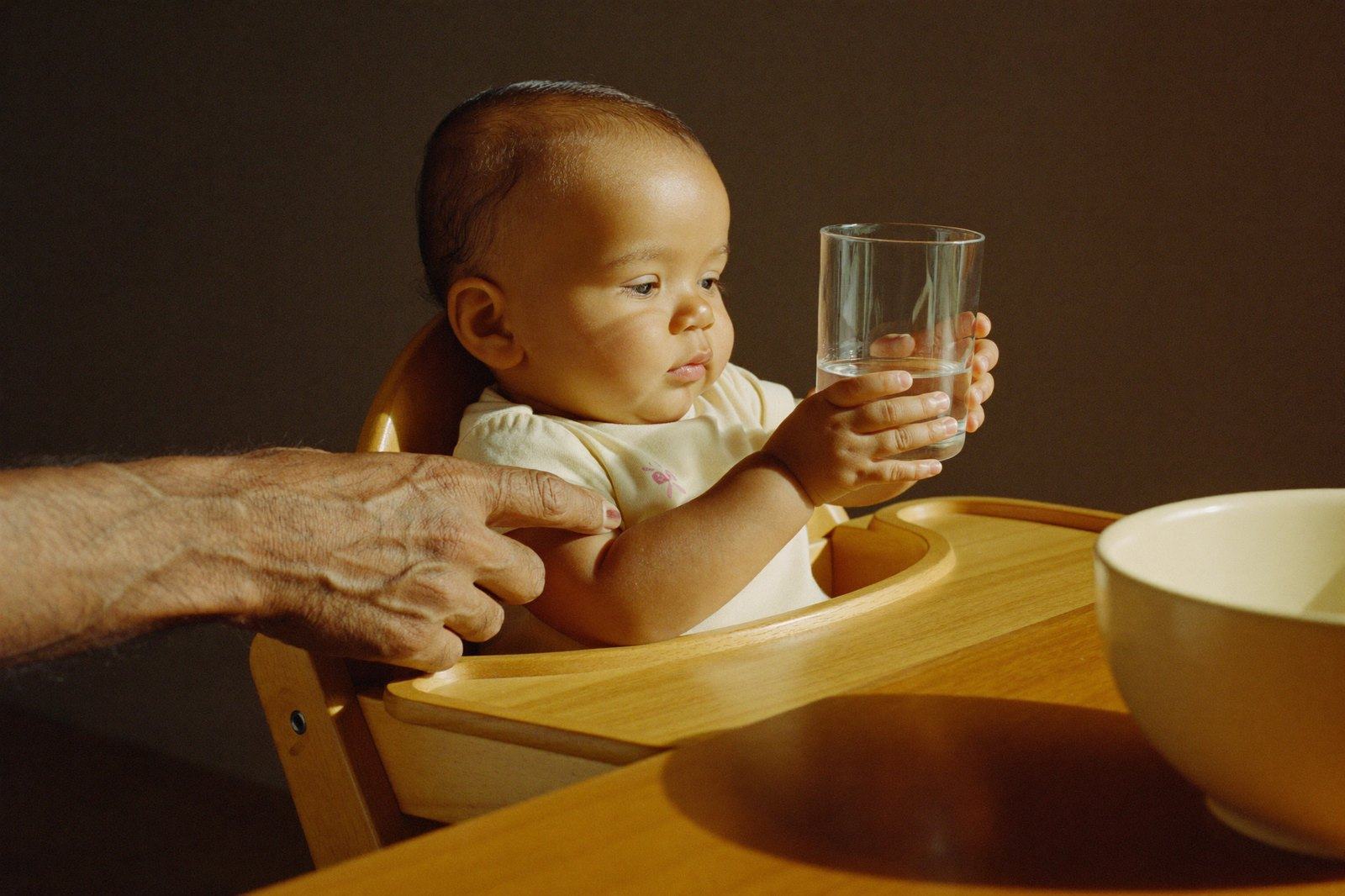 Un bebé en su periquera sostiene con las dos manos un vaso de vidrio pequeño con agua, con la mano de un adulto acompañándolo y un tazón al lado