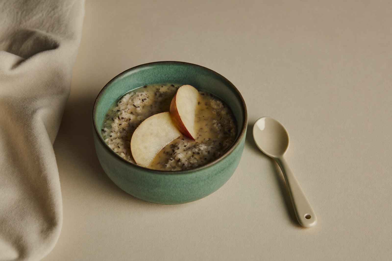 Tazón de cerámica verde con papilla de avena y rodajas de manzana, junto a una cuchara de bebé color crema sobre una mesa clara