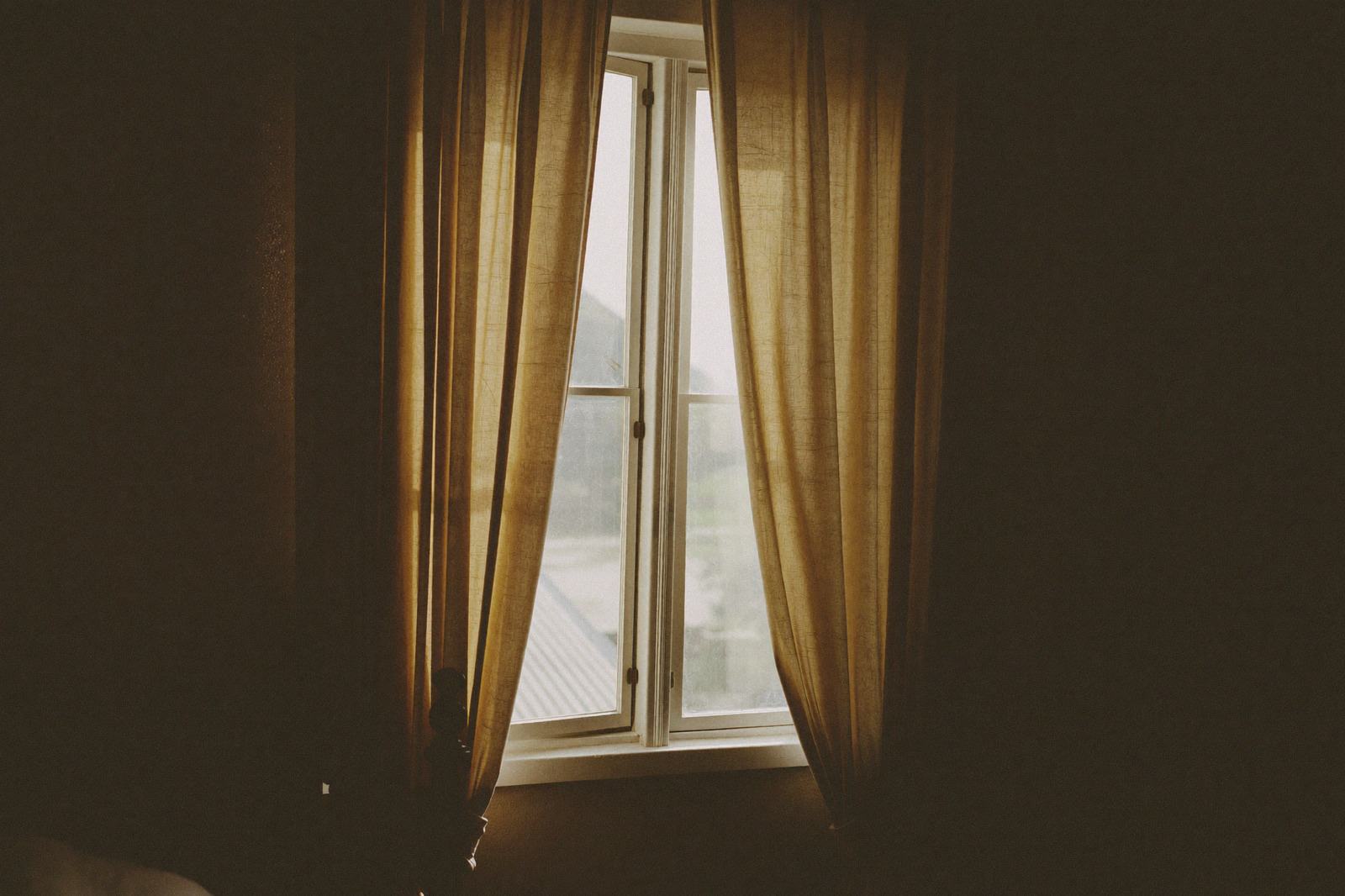 Ventana de una habitación en penumbra con las cortinas entrecerradas filtrando la luz del mediodía