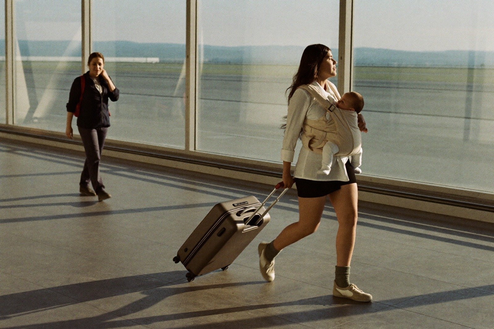 Mamá cruzando la sala de un aeropuerto con su bebé en el portabebés y una maleta de mano