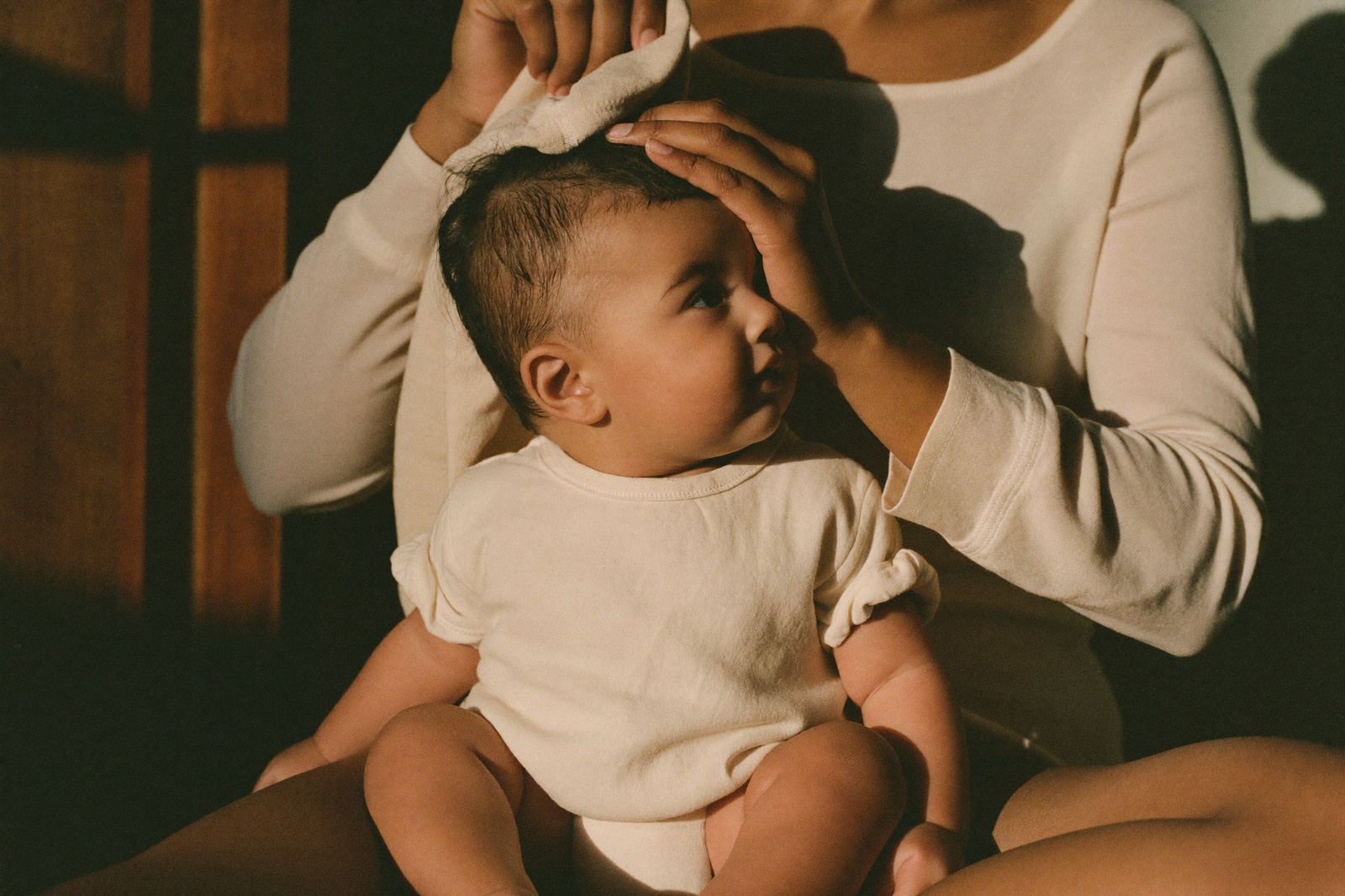 Madre secando con una toalla el cabello de su bebé, sentado en su regazo con la luz cálida de la tarde
