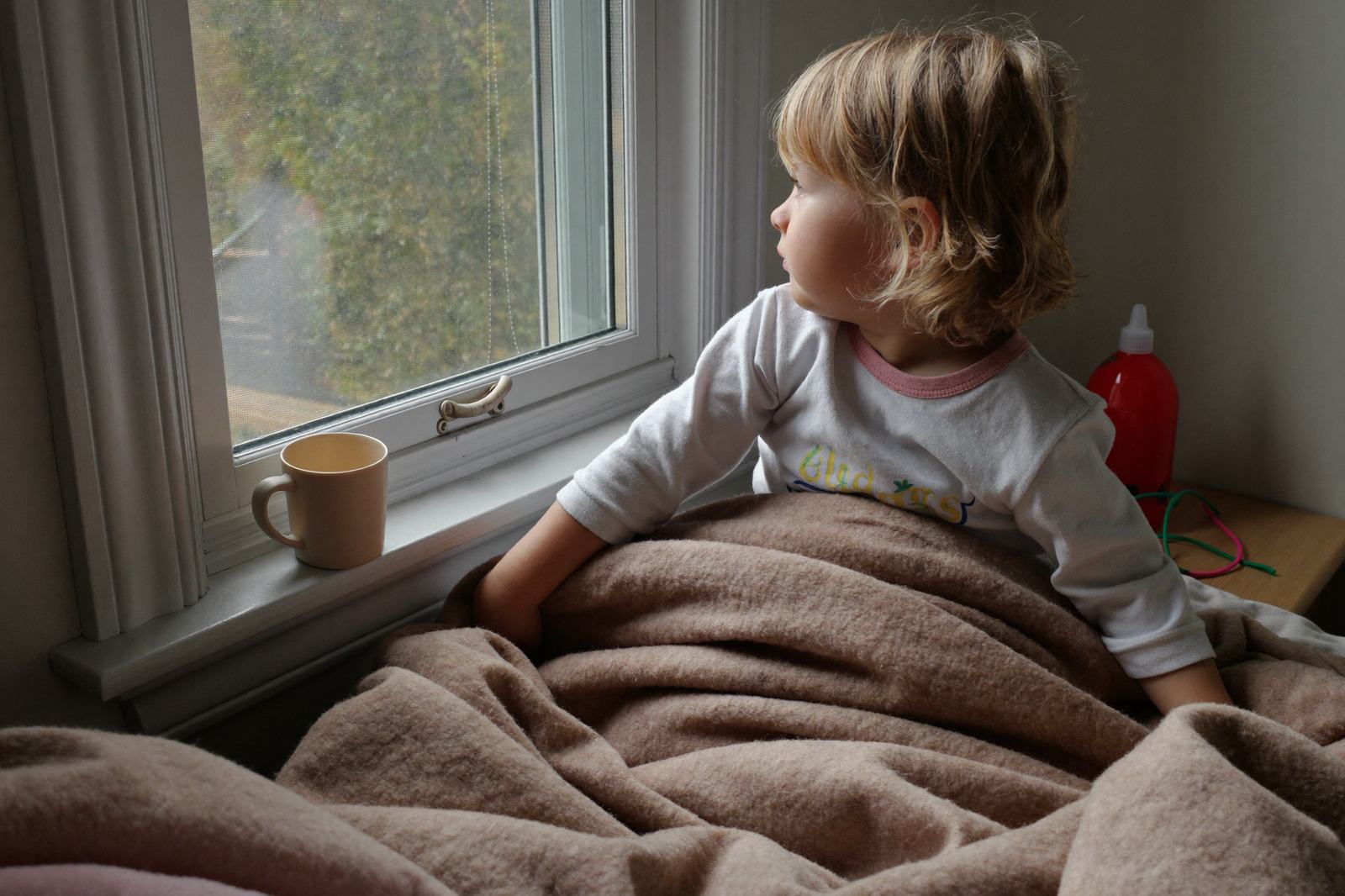 Niño pequeño en pijama sentado en la cama con una manta, mirando por la ventana