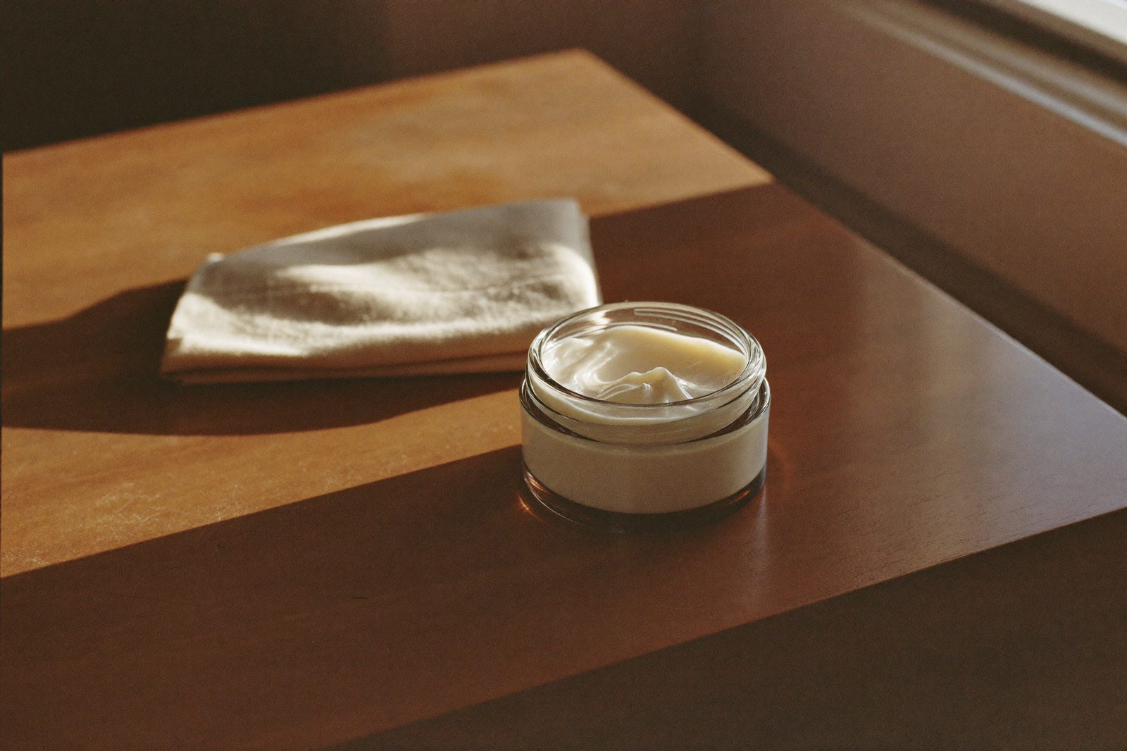 Frasco de crema sin etiqueta y una toalla doblada sobre una cómoda junto a una ventana