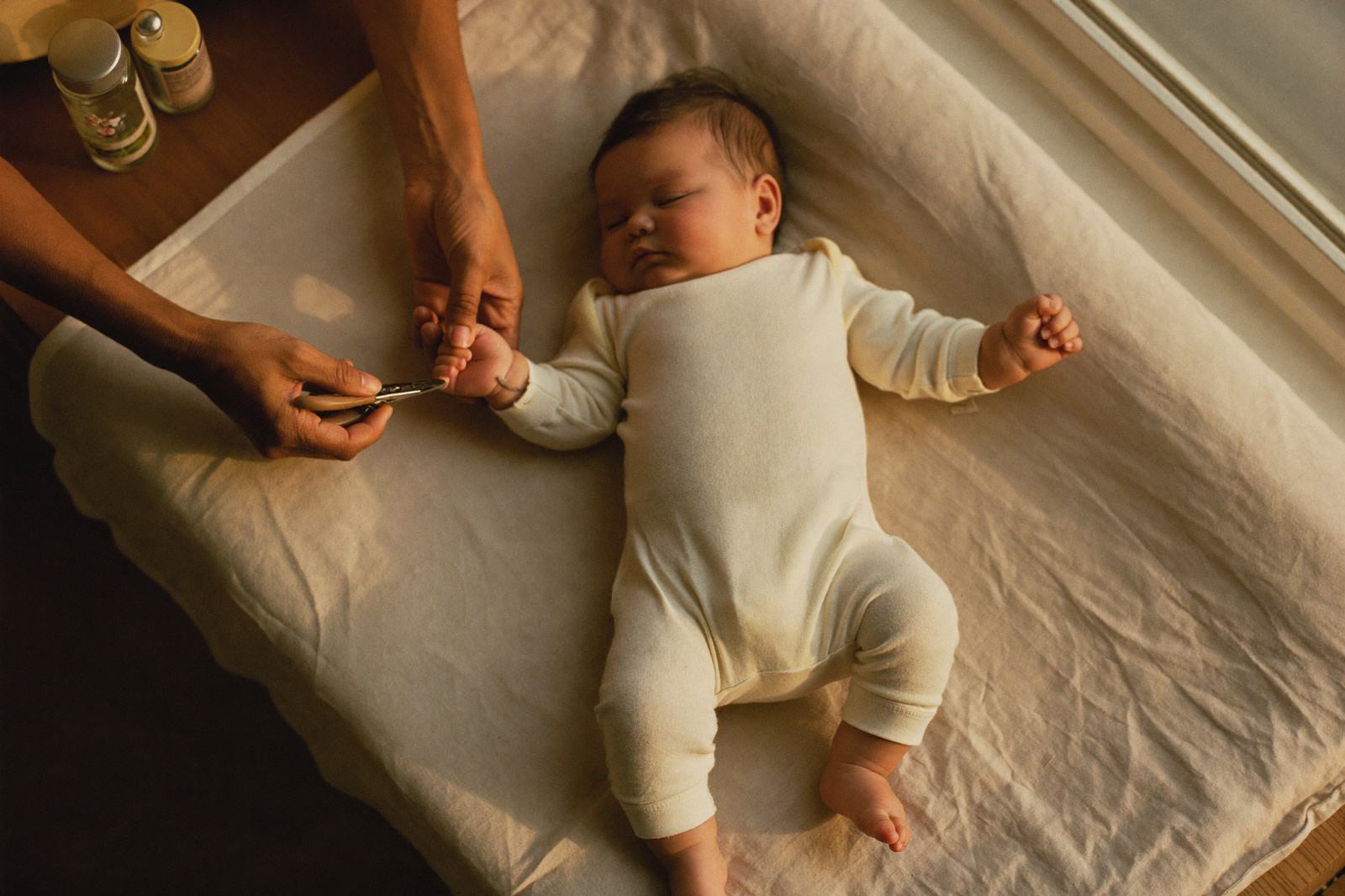 Manos de una madre sosteniendo la manita de su bebé dormido para cortarle las uñas junto a una ventana con luz suave