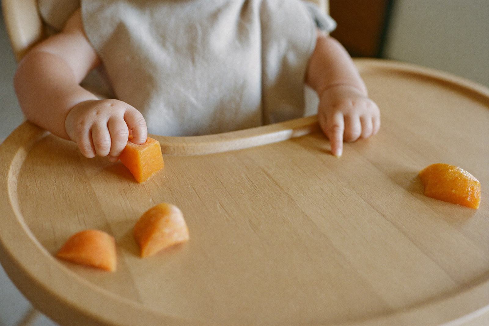 Bebé de nueve meses sentado en su silla alta acercando con los dedos un trozo de comida sobre la charola