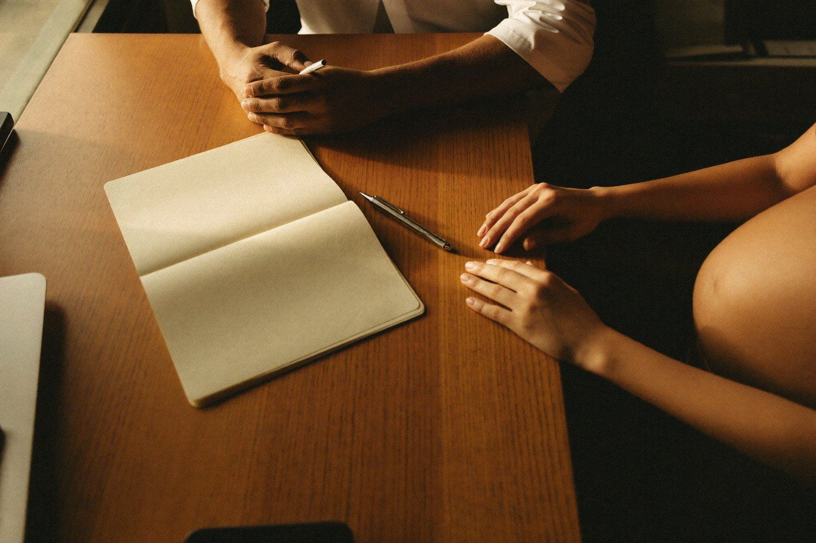 Vista superior del escritorio de un consultorio: una libreta abierta, la mano del personal de salud con una pluma y las manos de una mujer embarazada apoyadas en la mesa