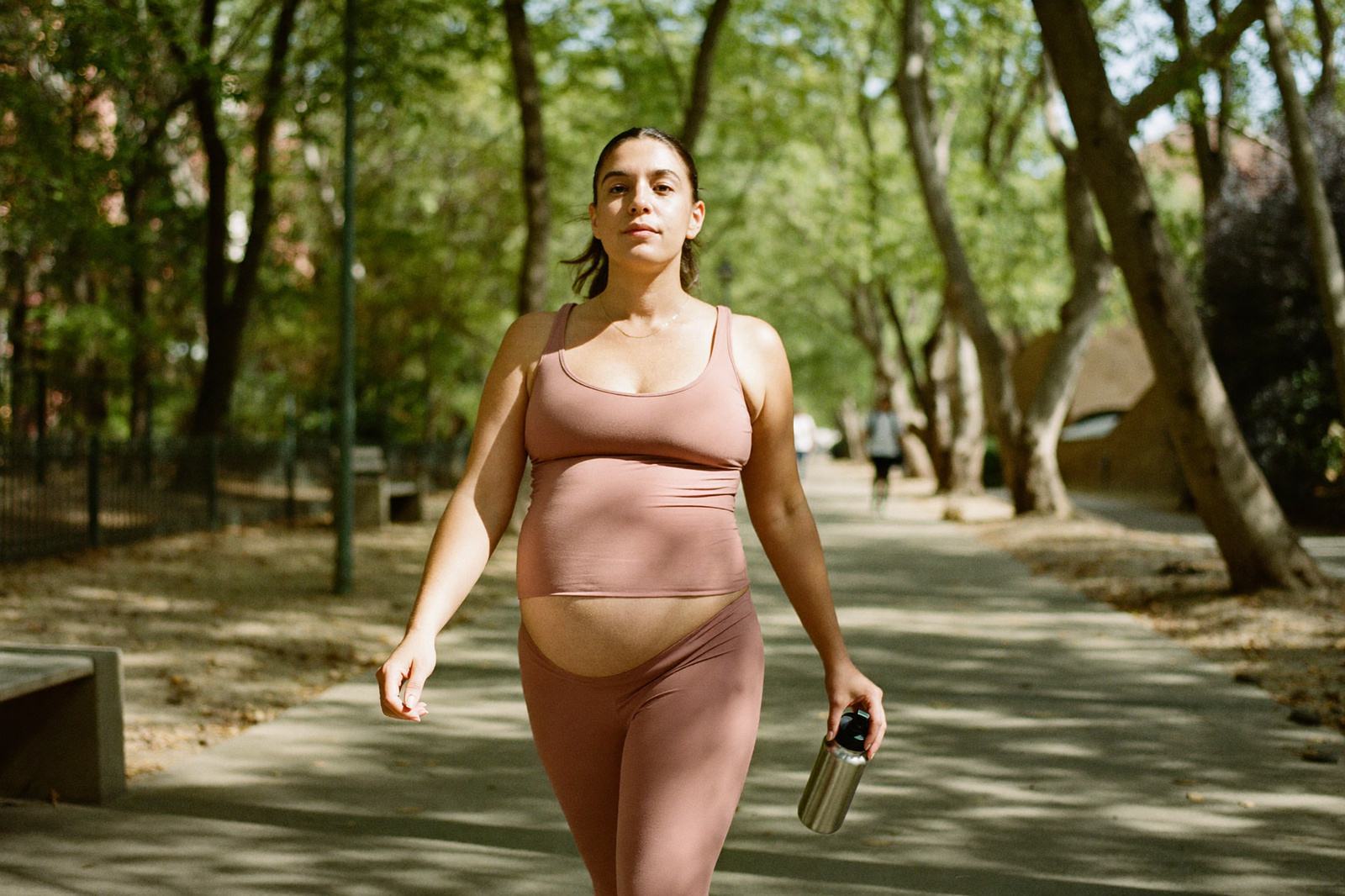 Mujer embarazada caminando por un parque arbolado a paso firme con ropa deportiva y una botella de agua