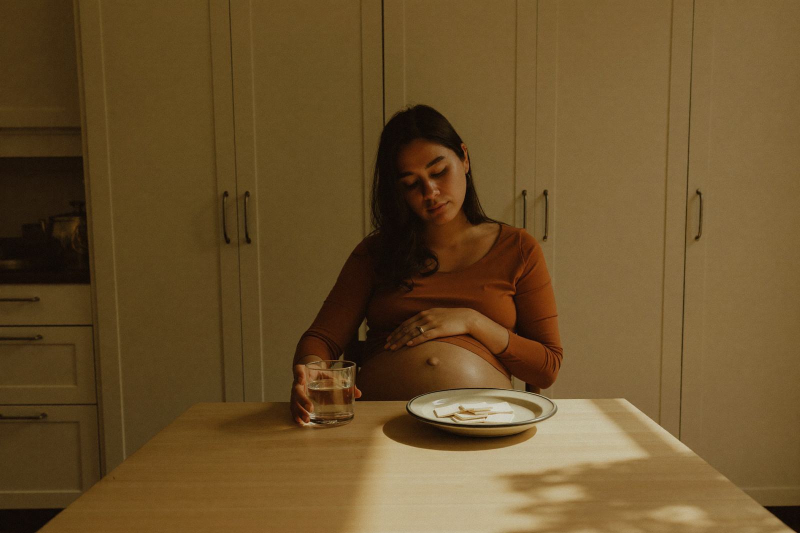 Mujer embarazada sentada a la mesa de su cocina por la tarde con un vaso y unas galletas saladas en un plato