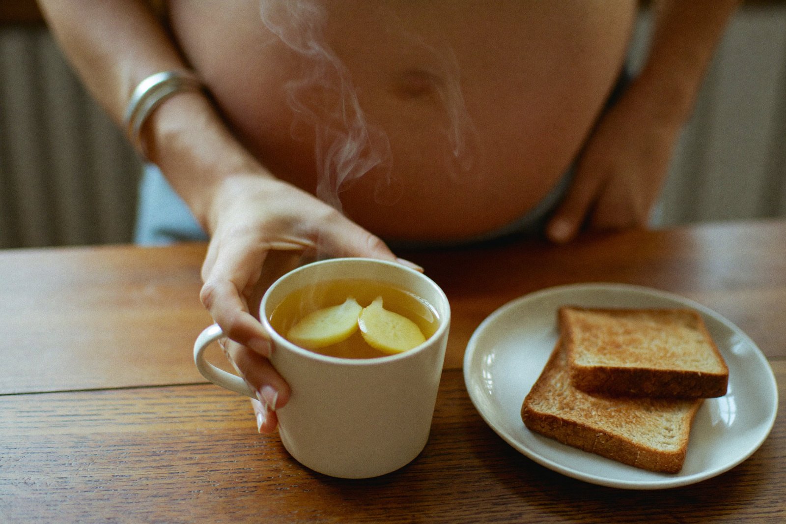 Manos de mujer embarazada sosteniendo una taza de té caliente con limón junto a un plato con dos rebanadas de pan tostado