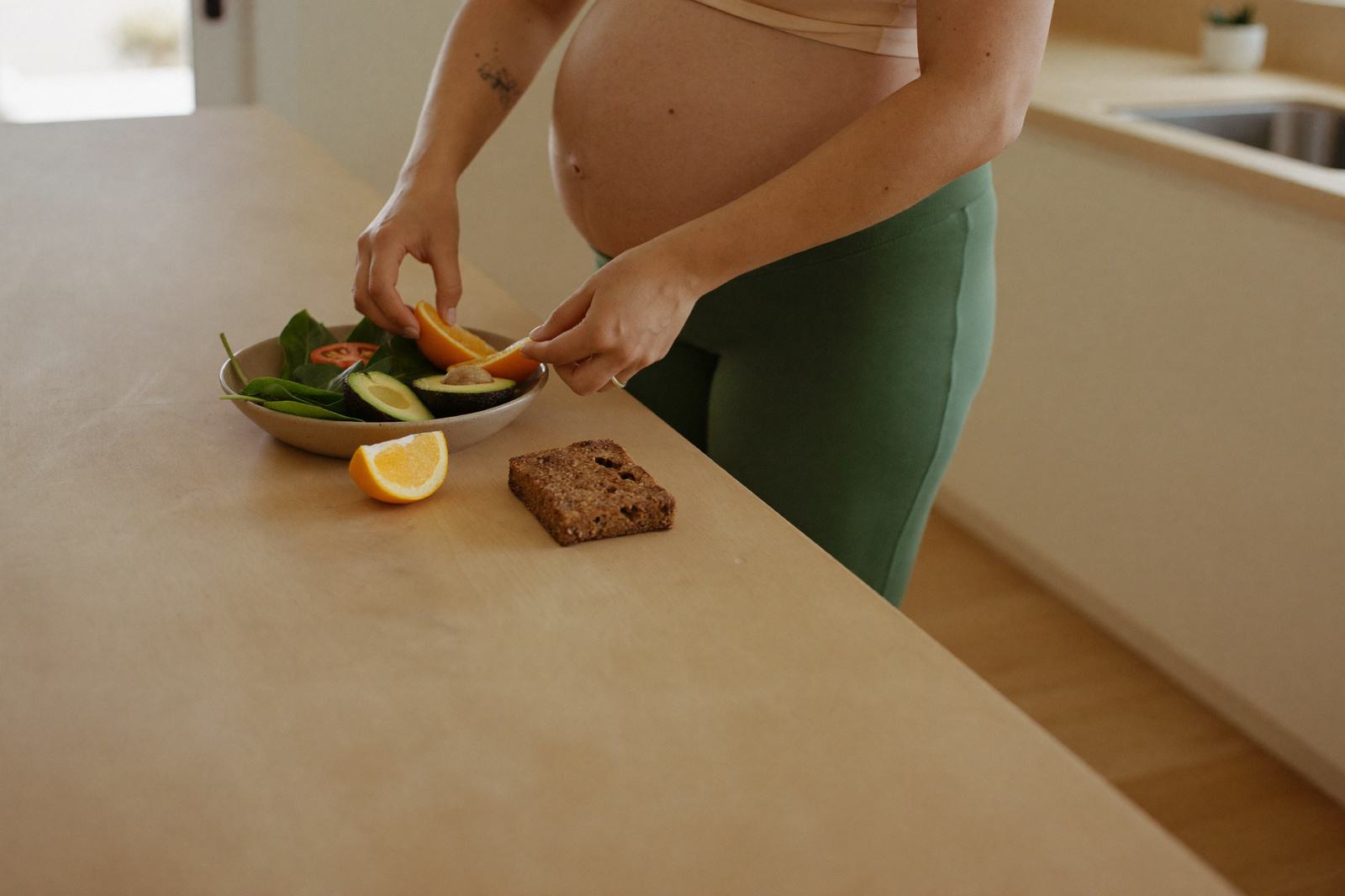 Mujer embarazada preparando un plato con verduras, fruta y pan integral sobre la barra de la cocina