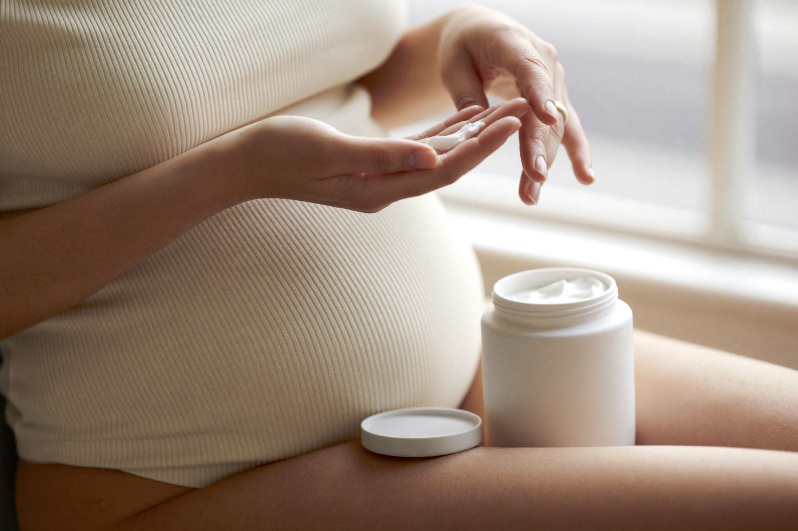 Mujer embarazada tomando crema de un frasco blanco abierto, sentada junto a una ventana
