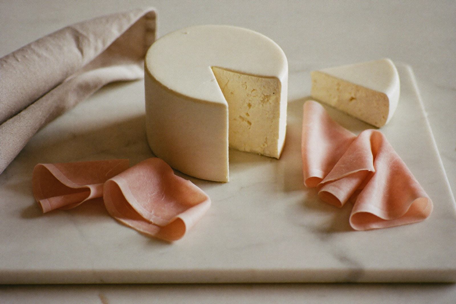 Queso fresco blanco y rebanadas delgadas de fiambre acomodadas sobre una tabla de mármol con una servilleta de lino, sin que nadie las toque