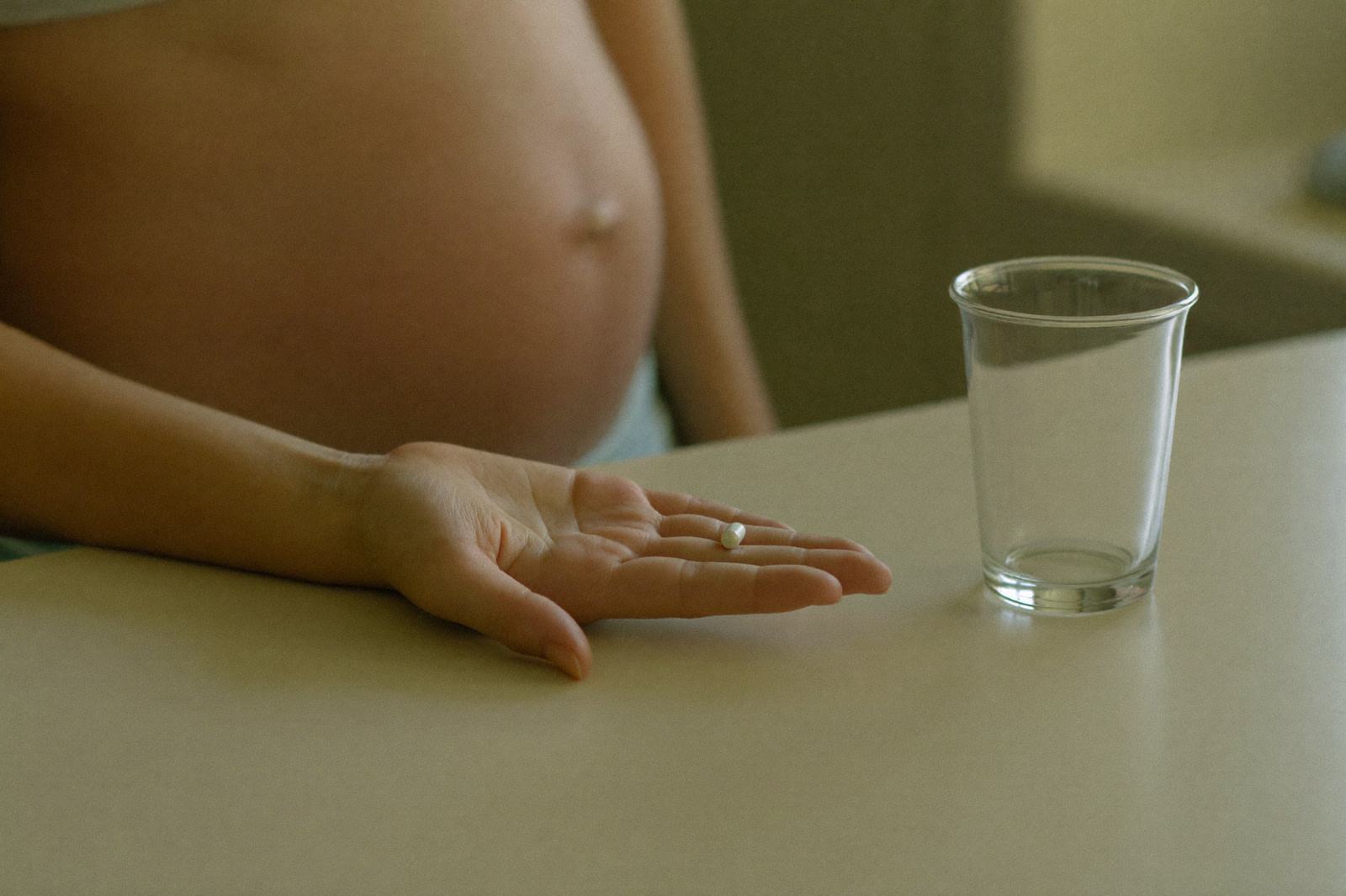 Mujer embarazada con una tableta de vitaminas prenatales en la palma de la mano junto a un vaso de agua sobre la mesa