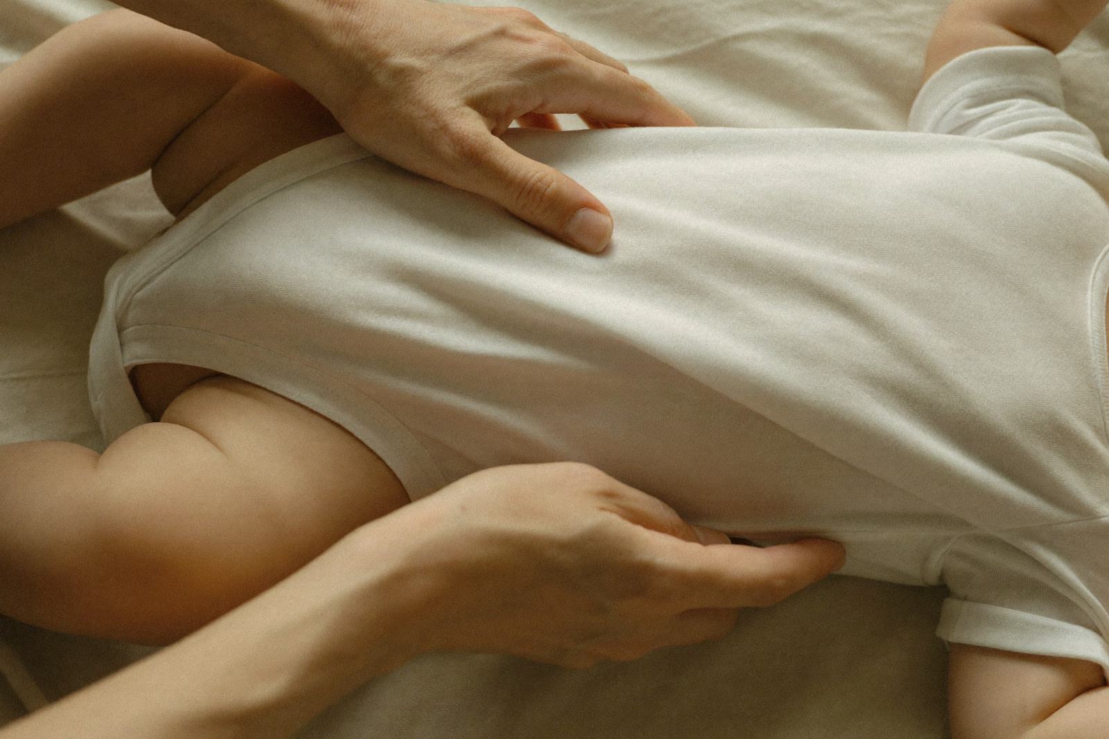 Manos de un adulto acomodando la ropa sobre el vientre de un bebé recostado, con luz suave de ventana