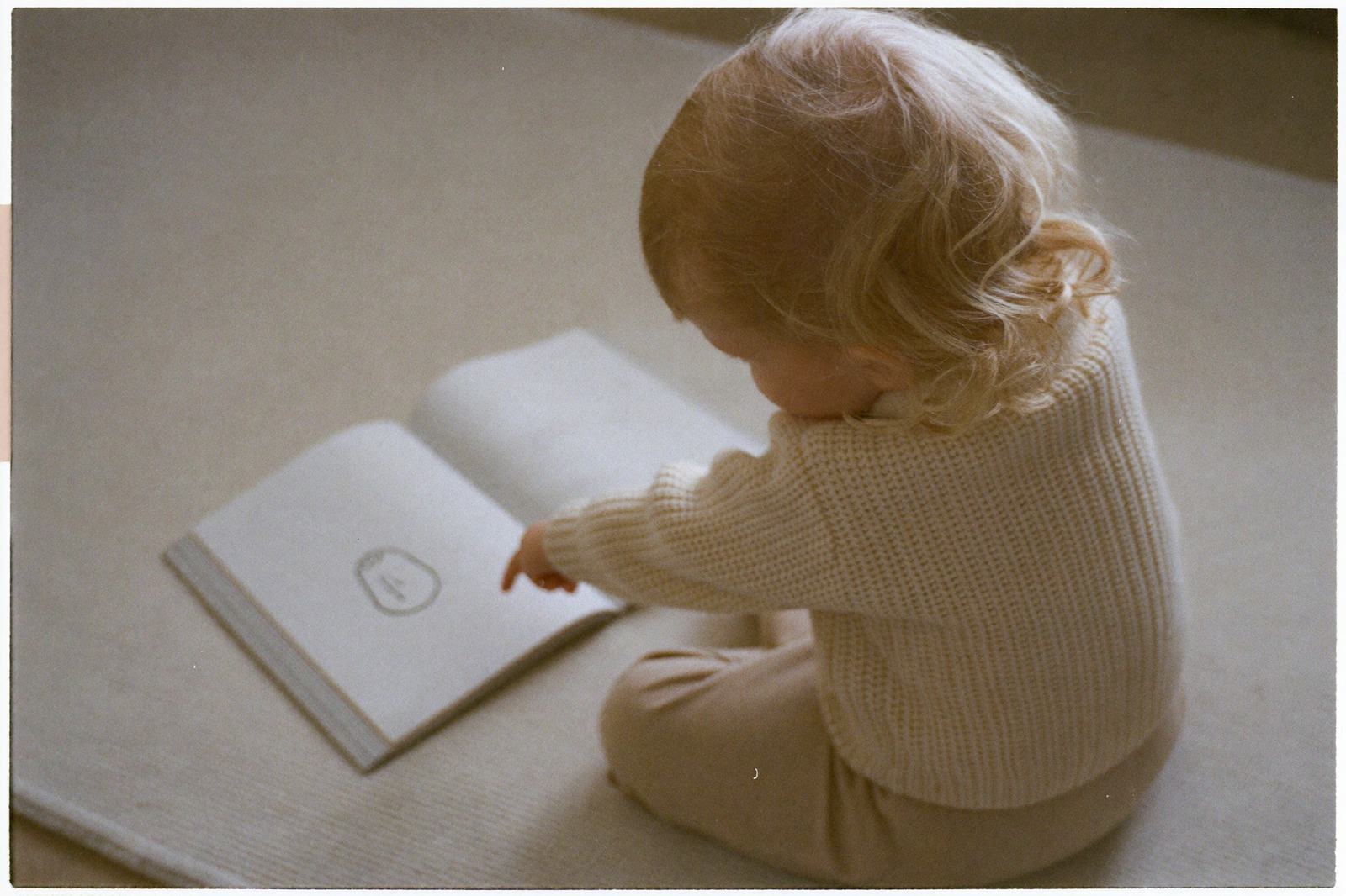Un niño de dos años sentado en el piso señalando una ilustración en un libro abierto sobre sus piernas