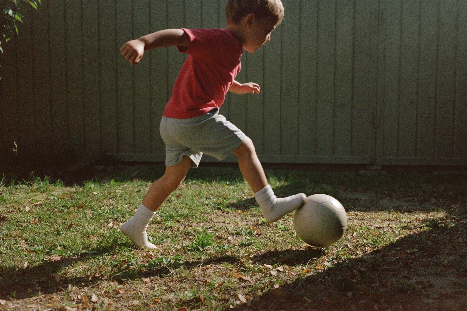 Un niño de dos años pateando una pelota en un patio con luz de tarde, visto de perfil