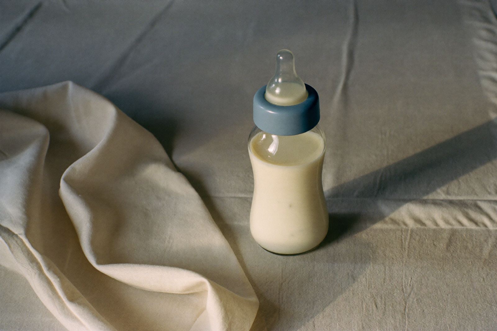 Un biberón de vidrio con leche de fórmula preparada, tapado, sobre un mantel de lino claro junto a una muselina doblada