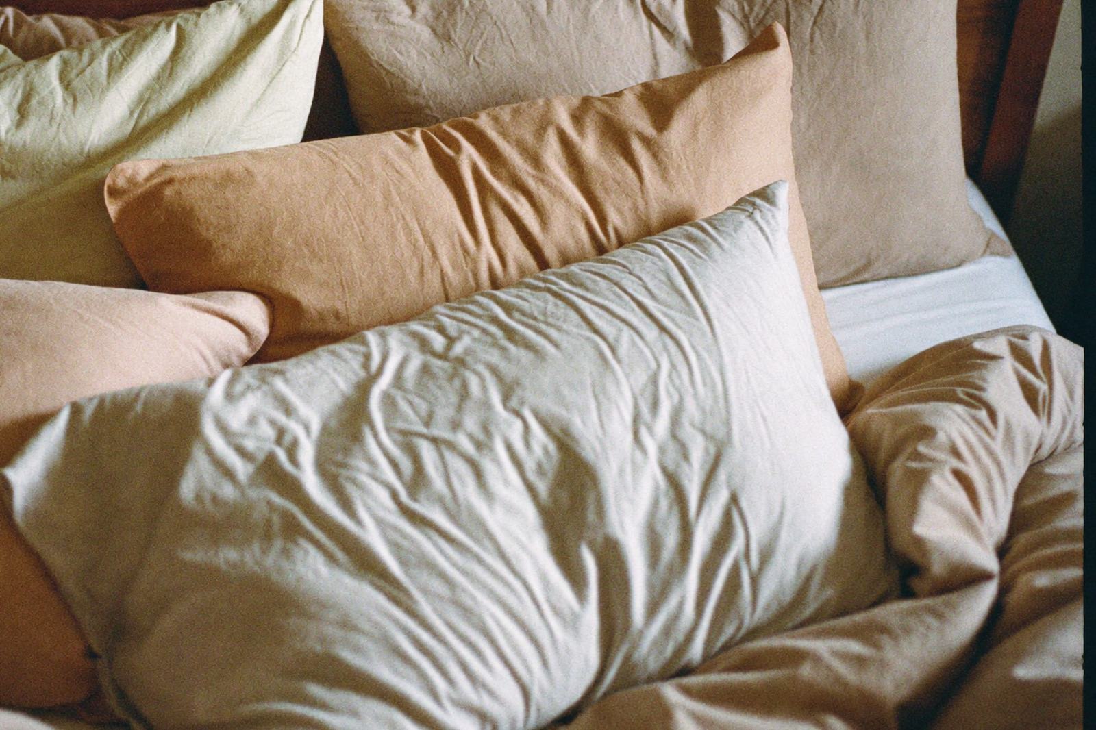 Almohadas apiladas sobre una cama junto a una manta doblada, con luz suave de ventana