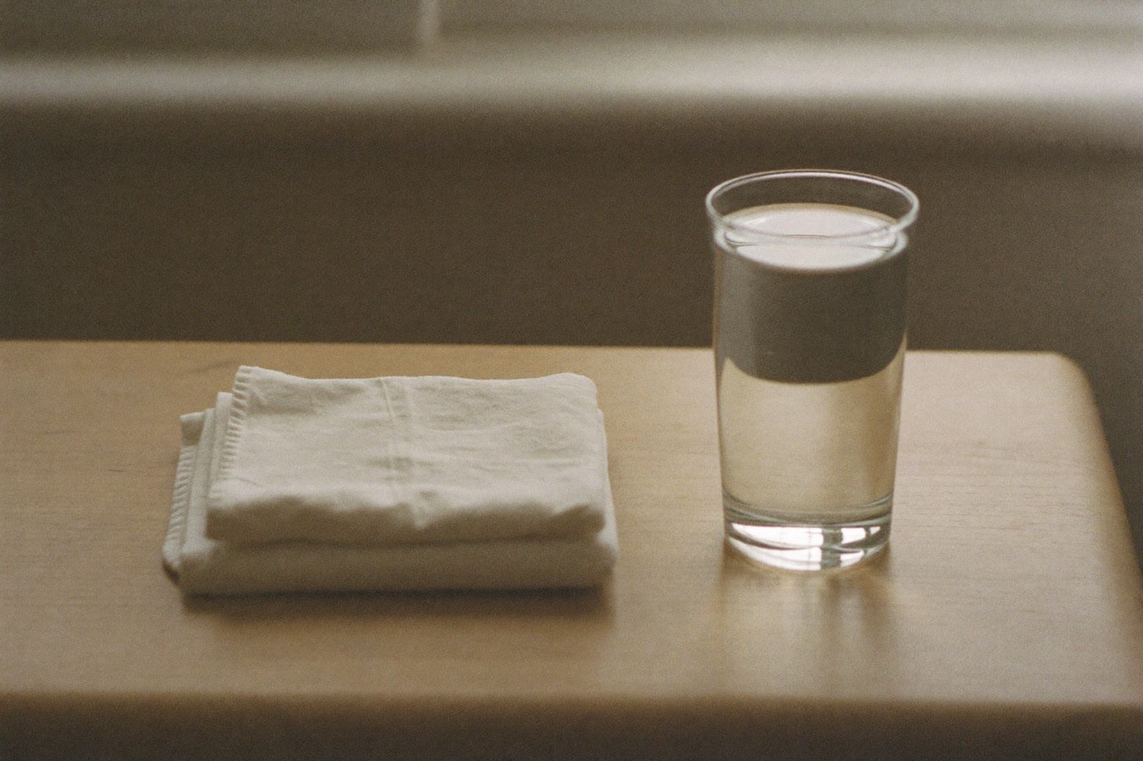 Una compresa de tela doblada junto a un vaso de agua sobre una mesa de madera clara, con luz de ventana