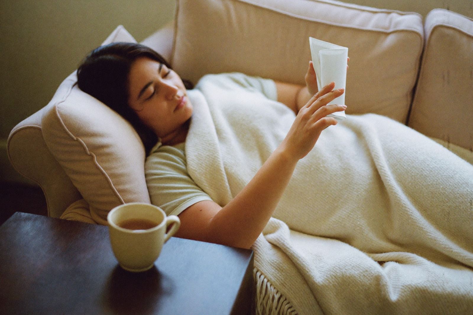 Una mamá recostada en un sillón bajo una manta, leyendo, con una taza caliente en la mesa lateral