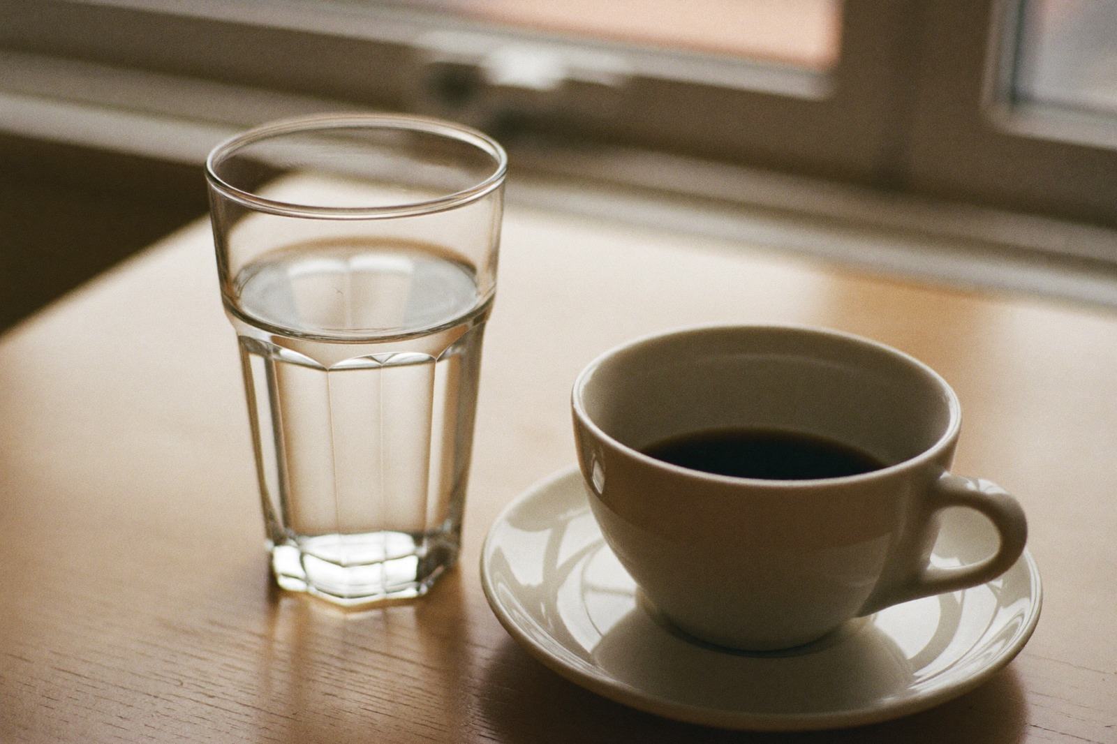 Un vaso de agua y una taza de café sobre una mesa junto a una ventana con luz de mañana